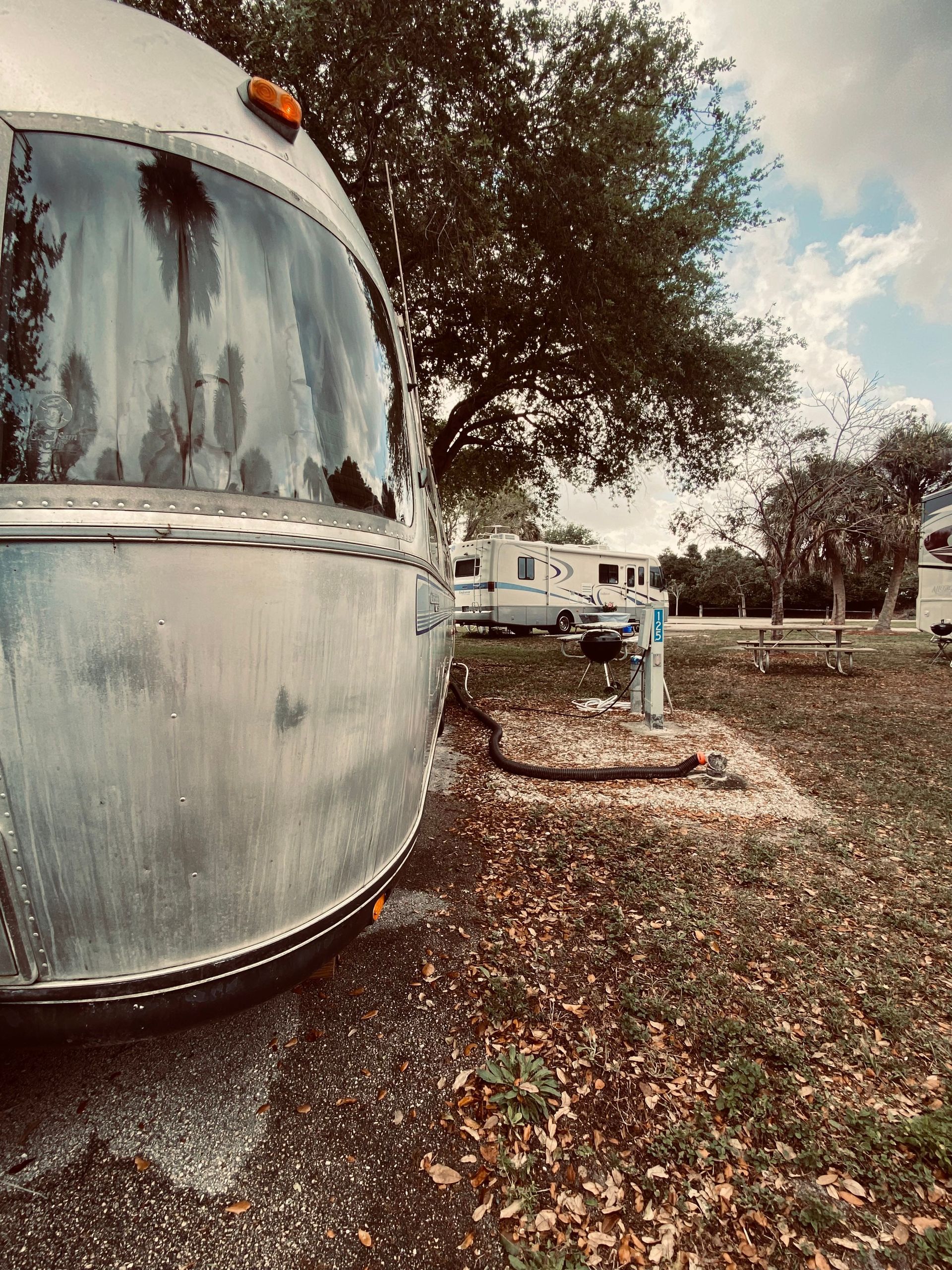 A side view of a silver Airstream camper parked at a campground with another RV visible in the background.