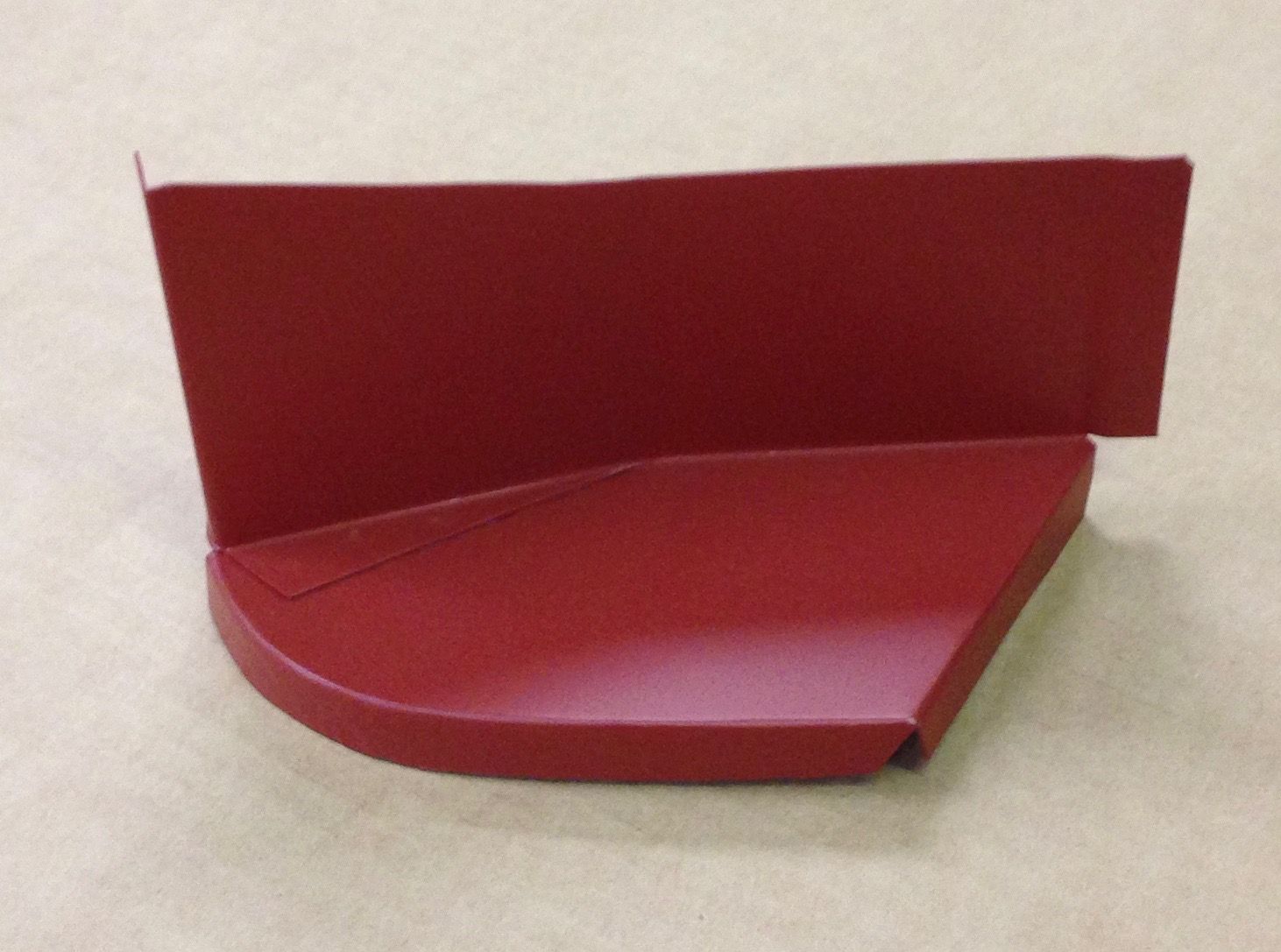 Red metal car part, with curved bottom and angled side, on a beige surface.