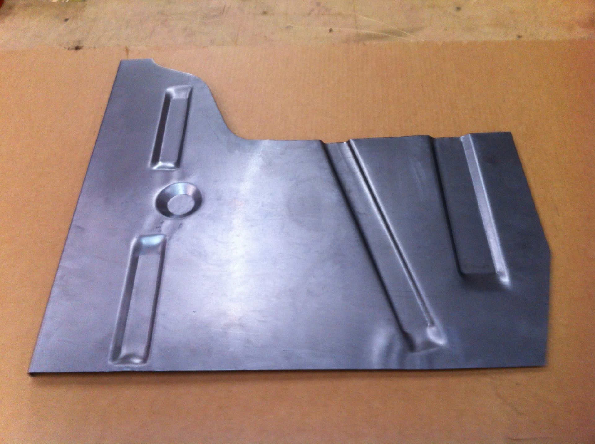 A gray metal car floor panel with raised sections and a circular hole on a wooden surface.