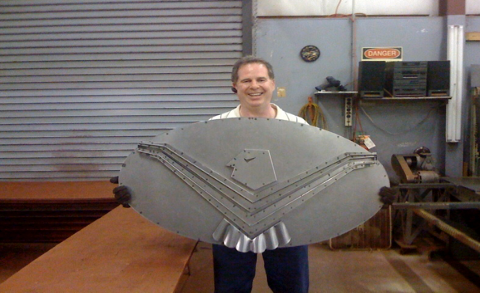 Man holding a large, gray metal shield with a bird emblem in a workshop; he smiles.