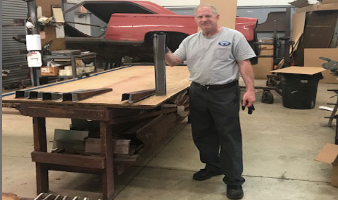 Man in shop with metal pieces on workbench, a car in background.