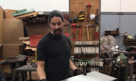 Man in shop holding metal sheet, tools and welding equipment in background.