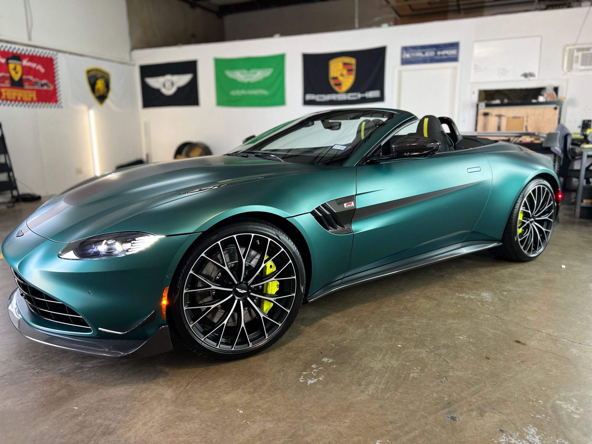 Green Aston Martin convertible parked inside a shop with car brand flags on the wall.