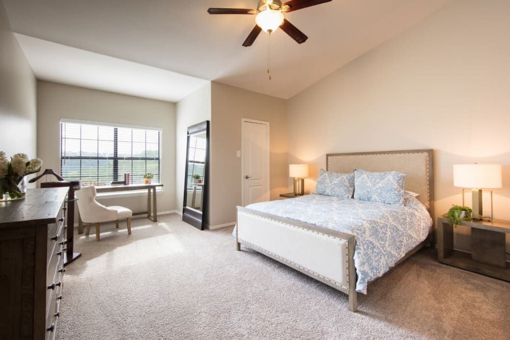 Apartment bedroom with a bed, dresser, chair, windows, and ceiling fan at Marquis at Treetops in Northwest Austin, TX.