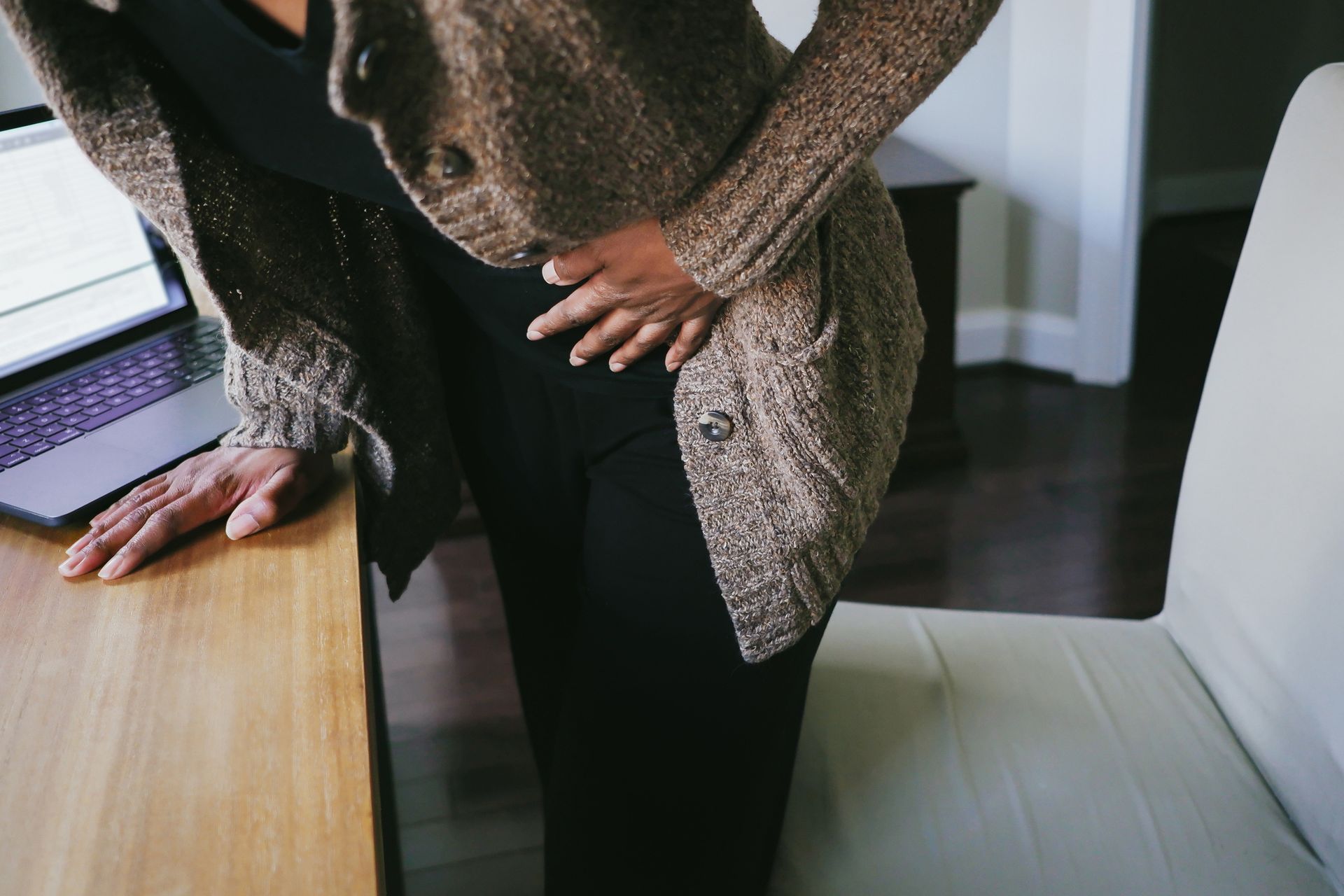 Woman clutching stomach, leaning on a desk with a laptop. Brown sweater, black pants, indoor setting.