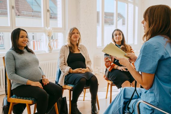 Four women, three pregnant, in a circle, one speaking while holding papers; bright room, white walls.