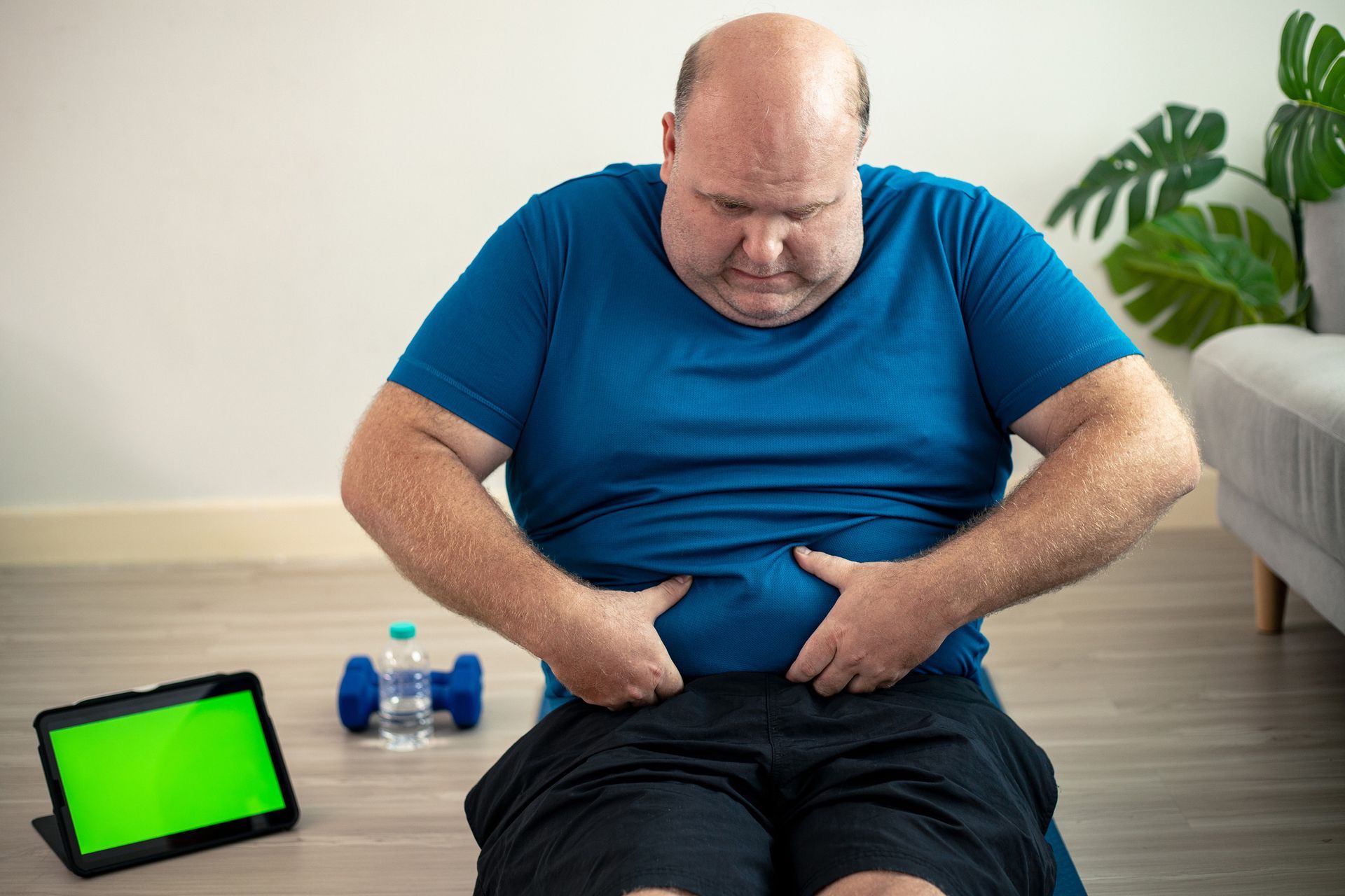 Man in blue shirt and black shorts, sitting, touching his stomach; a tablet, water bottle, and dumbbells nearby.