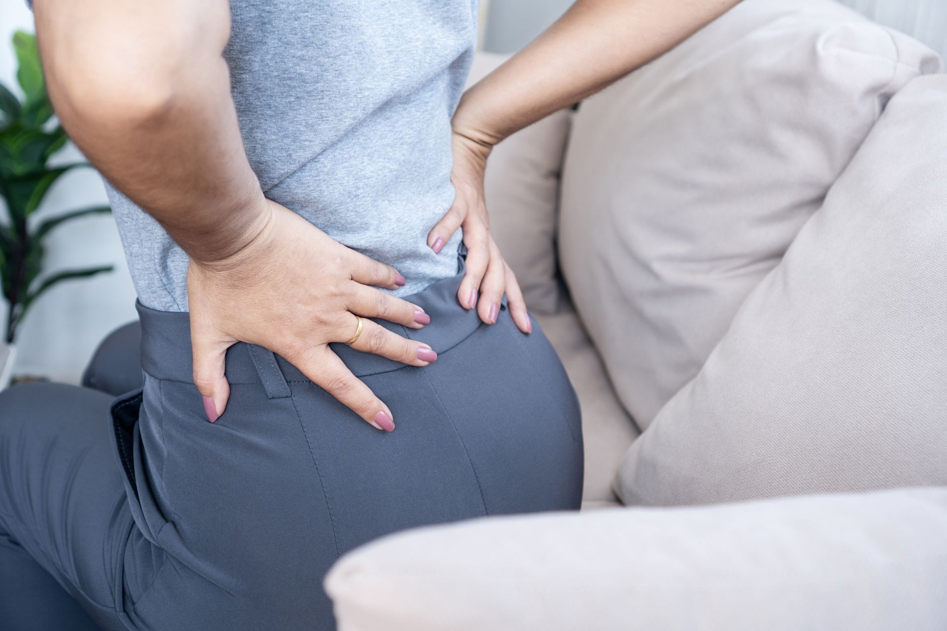 Woman holding her lower back, sitting on a sofa, possibly experiencing pain.