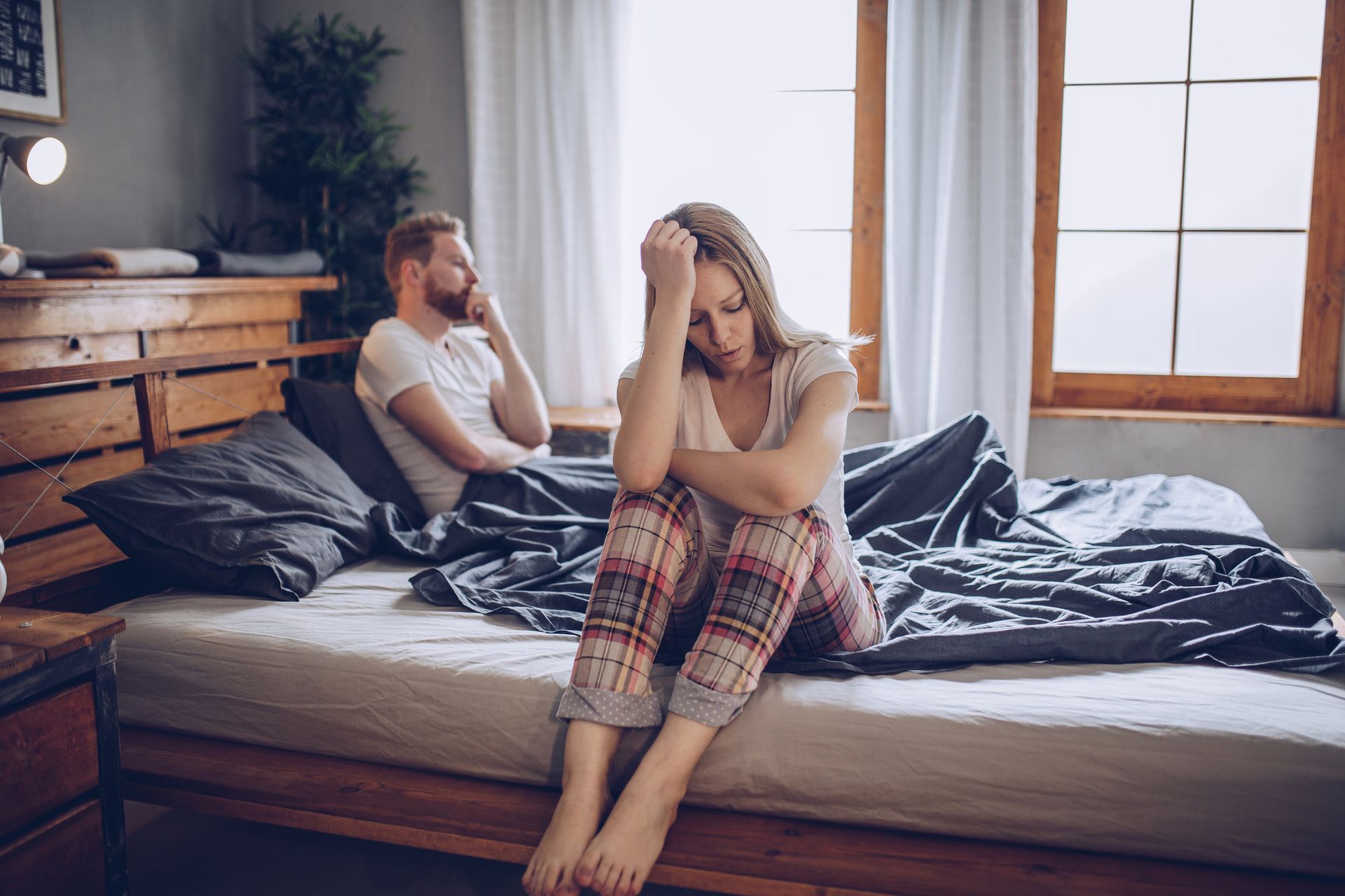 Woman with head in hands sitting on bed, man looking away, appears distressed, bedroom setting.