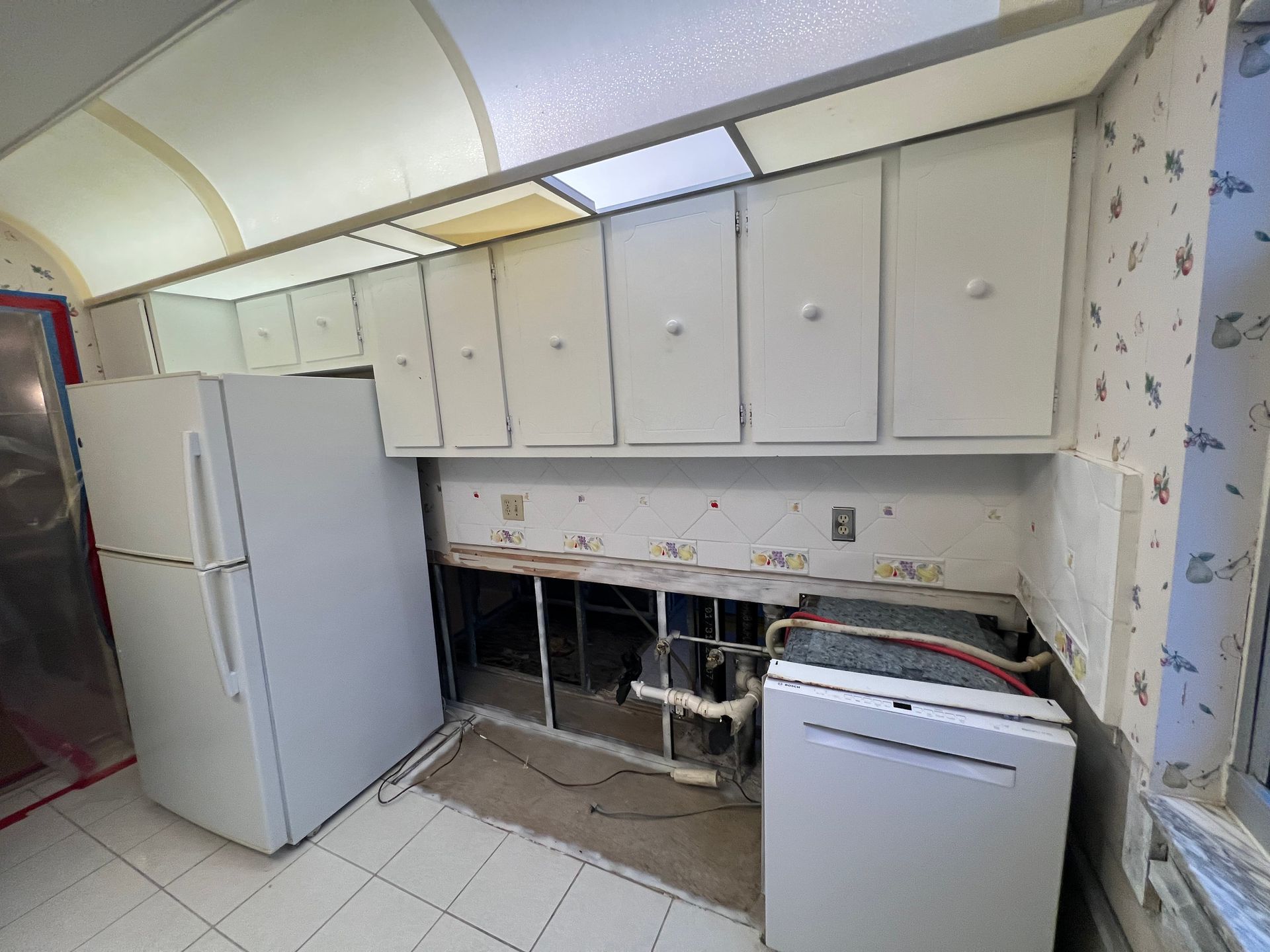Kitchen undergoing renovation with upper cabinets, fridge, and dishwasher. Walls are damaged.