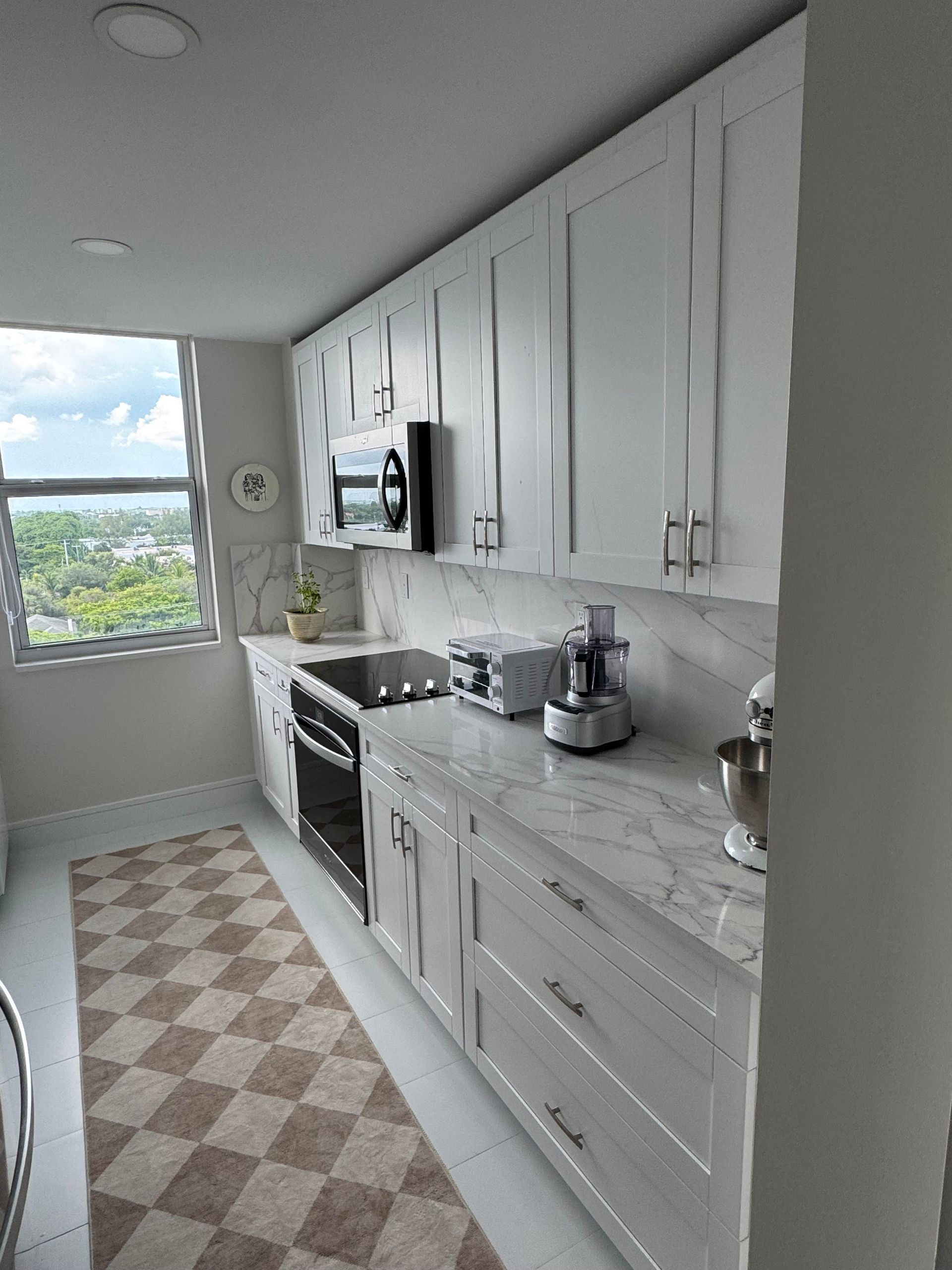 White kitchen with stainless steel refrigerator, white cabinets, checkered rug, and window with a view.