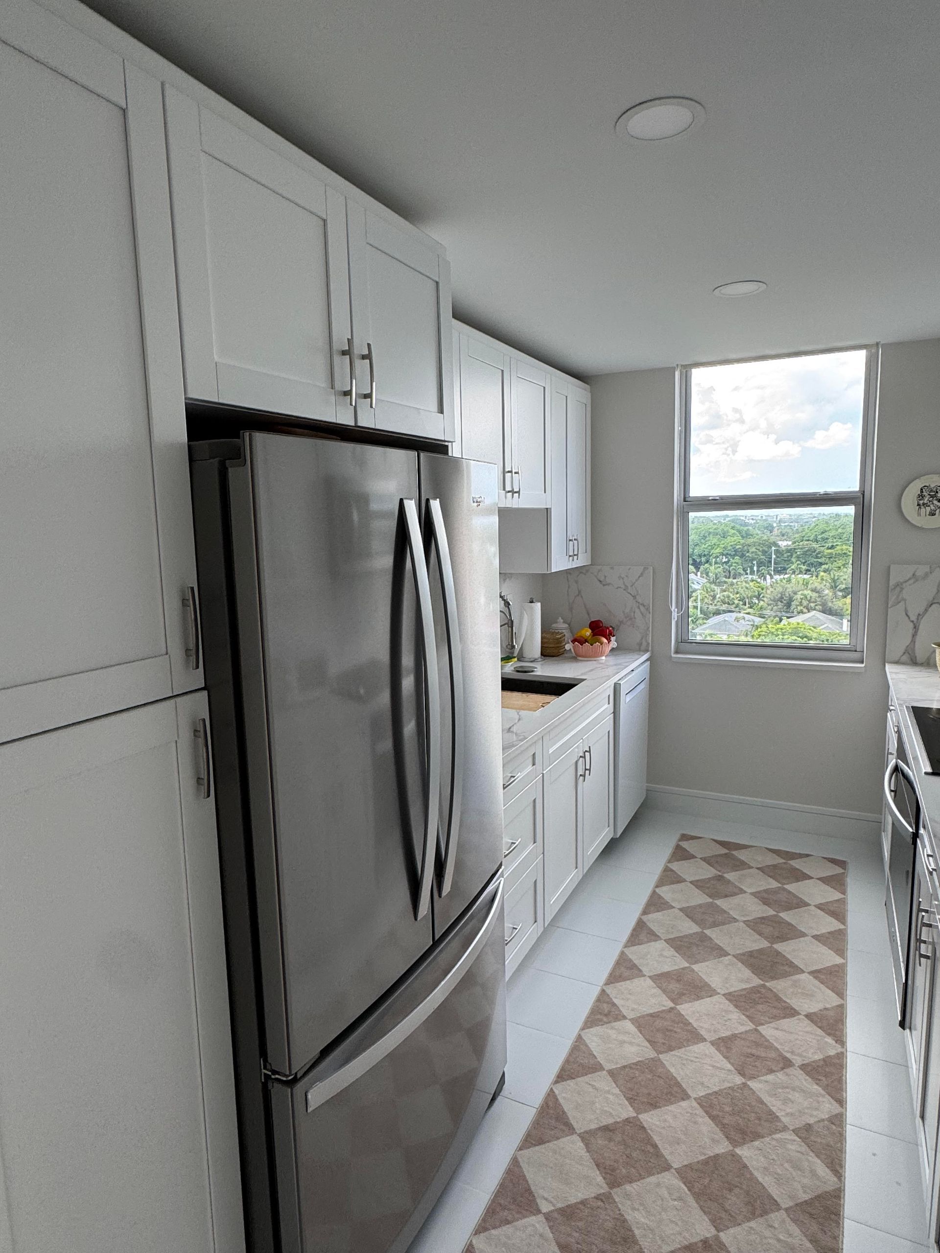 White kitchen with stainless steel refrigerator, white cabinets, checkered rug, and window with a view.