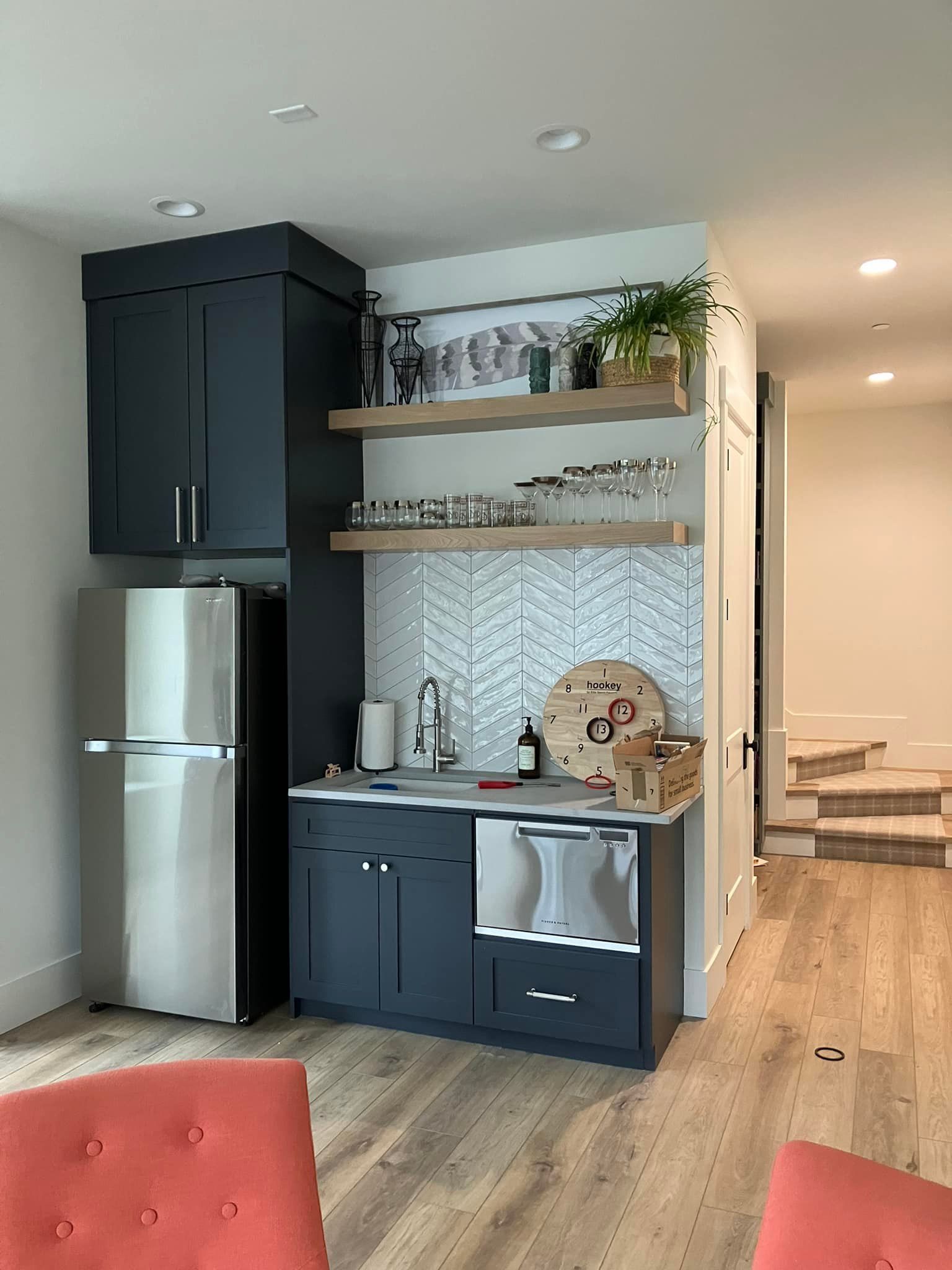 A navy wet bar with a stainless steel fridge, sink, and dishwasher, featuring open wood shelving and a patterned backsplash.
