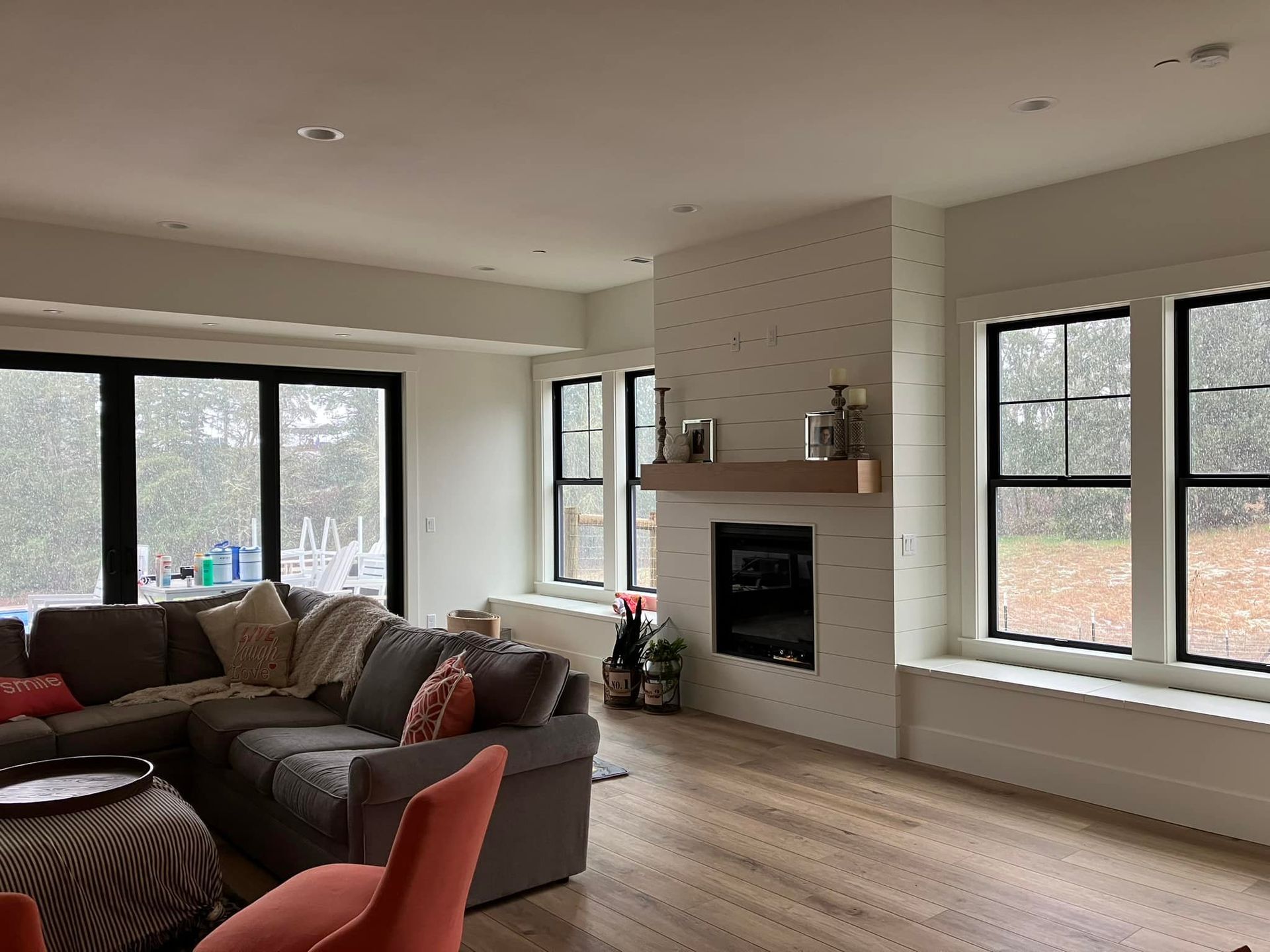 A cozy living room with a large gray sectional sofa, a fireplace with a wood mantel, and windows looking out at snow.