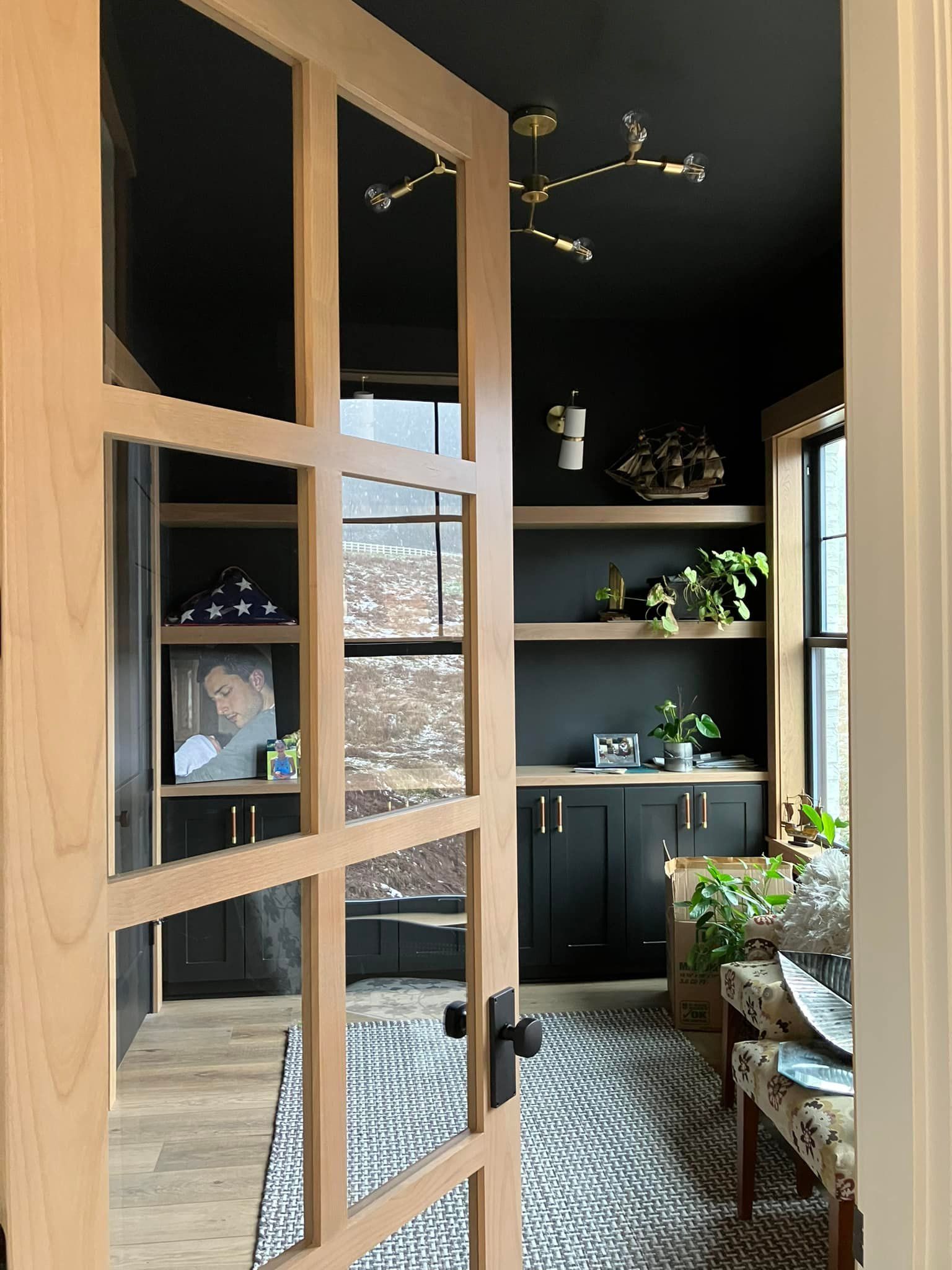 A view through a wooden French door into a modern office with black walls, a light fixture, shelving, and a patterned rug.
