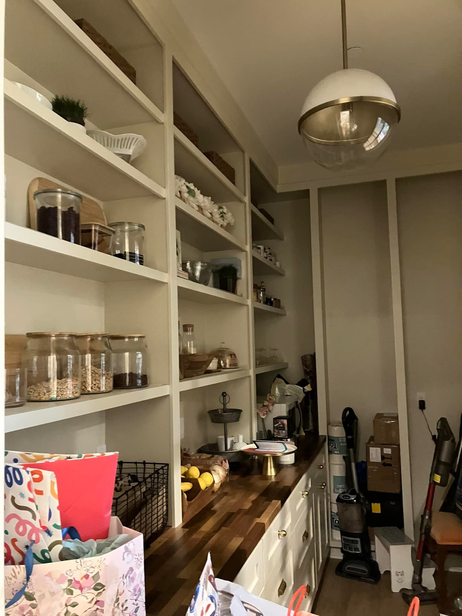 A walk-in pantry featuring white shelving filled with glass storage jars, a wooden countertop, and a modern globe pendant.