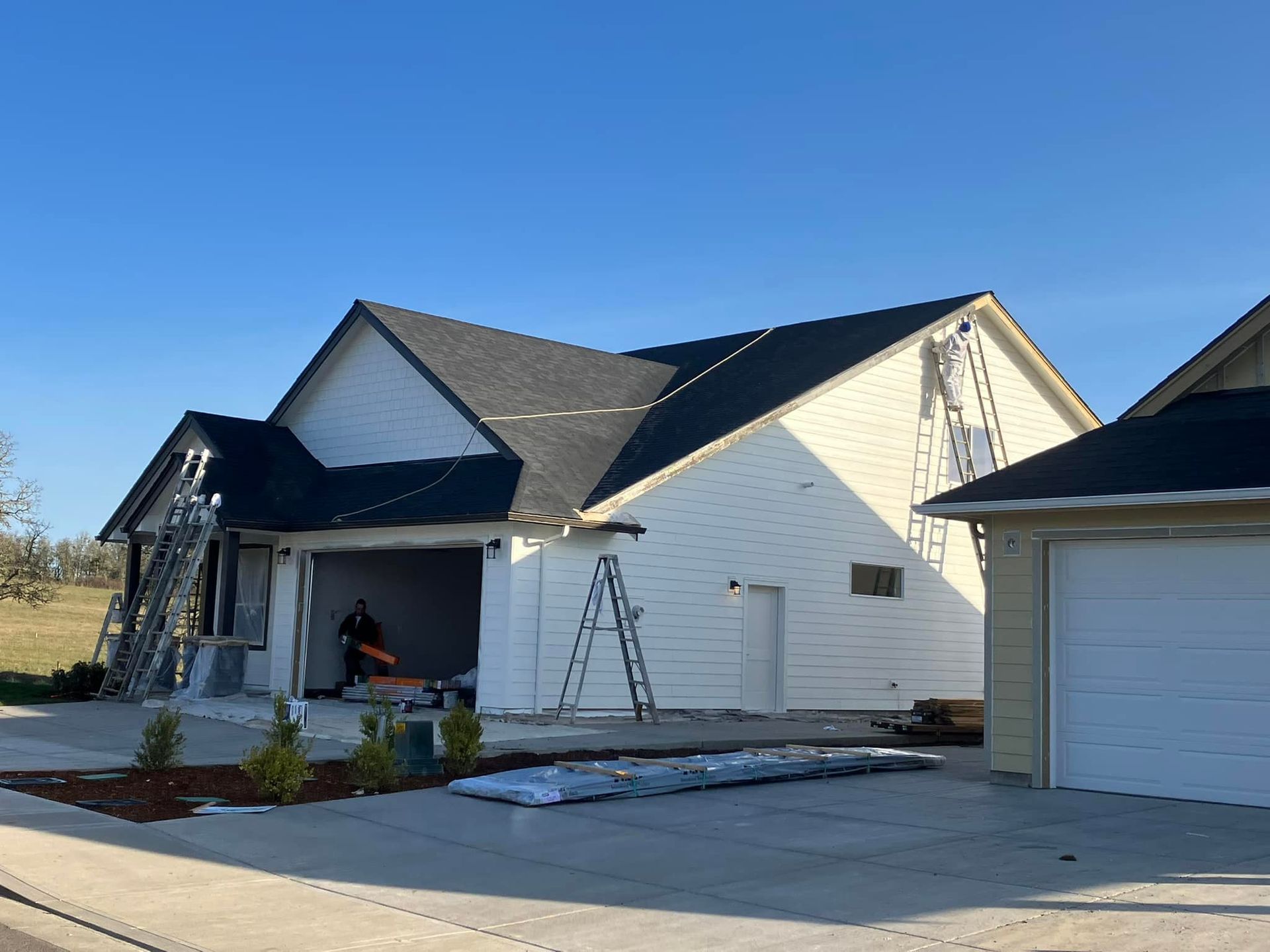 A white house under construction with a dark shingled roof, a worker in the garage, and ladders leaning against the siding.