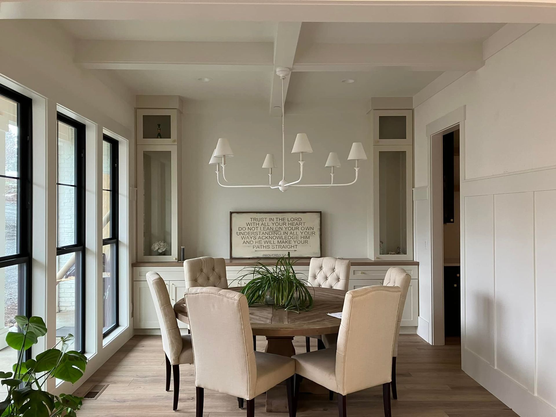 A dining room with a round wooden table, six beige upholstered chairs, a white multi-light chandelier, and glass cabinets.