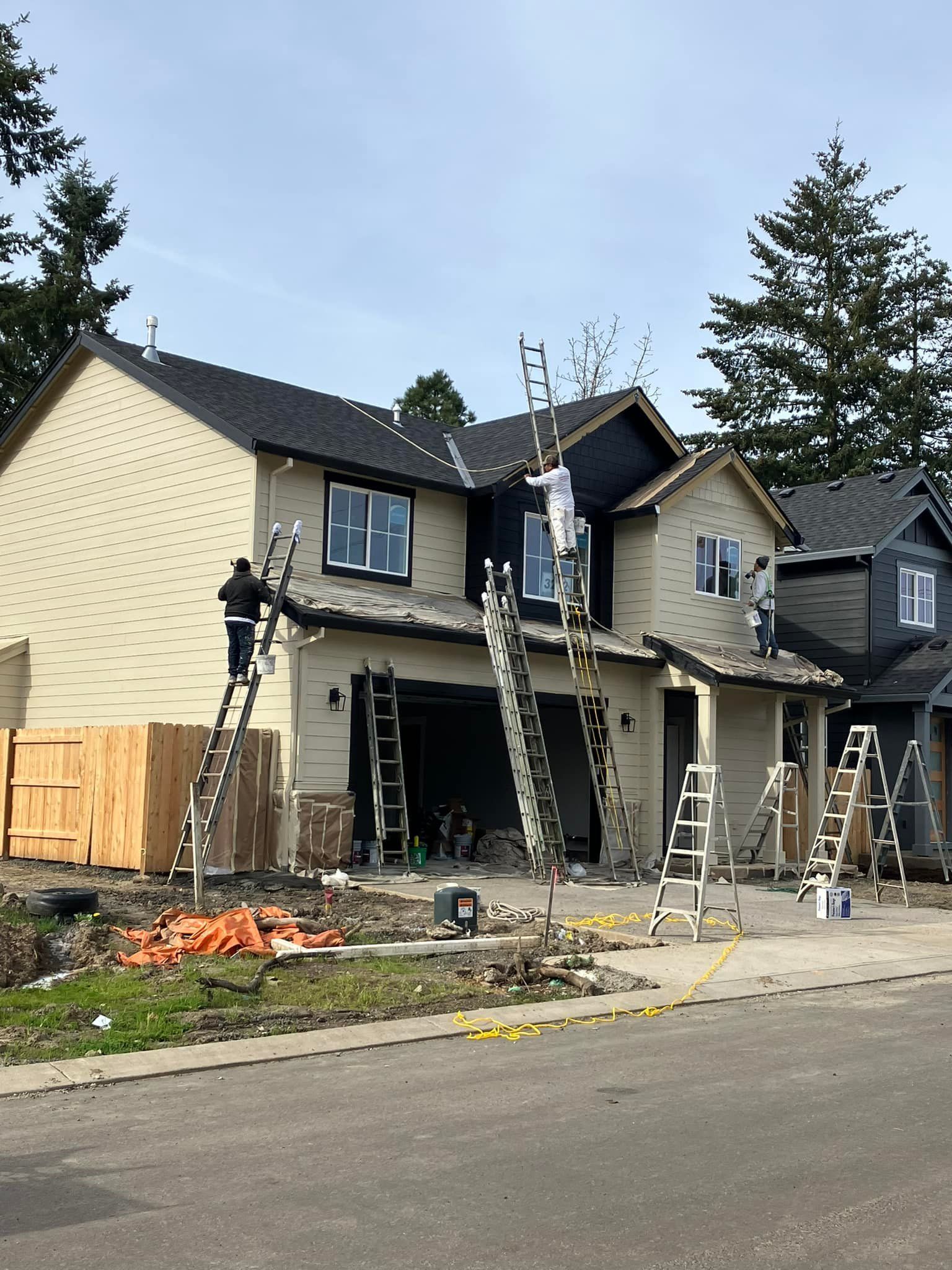 Construction workers on ladders working on the siding and roofing of a two-story beige house under a clear blue sky.