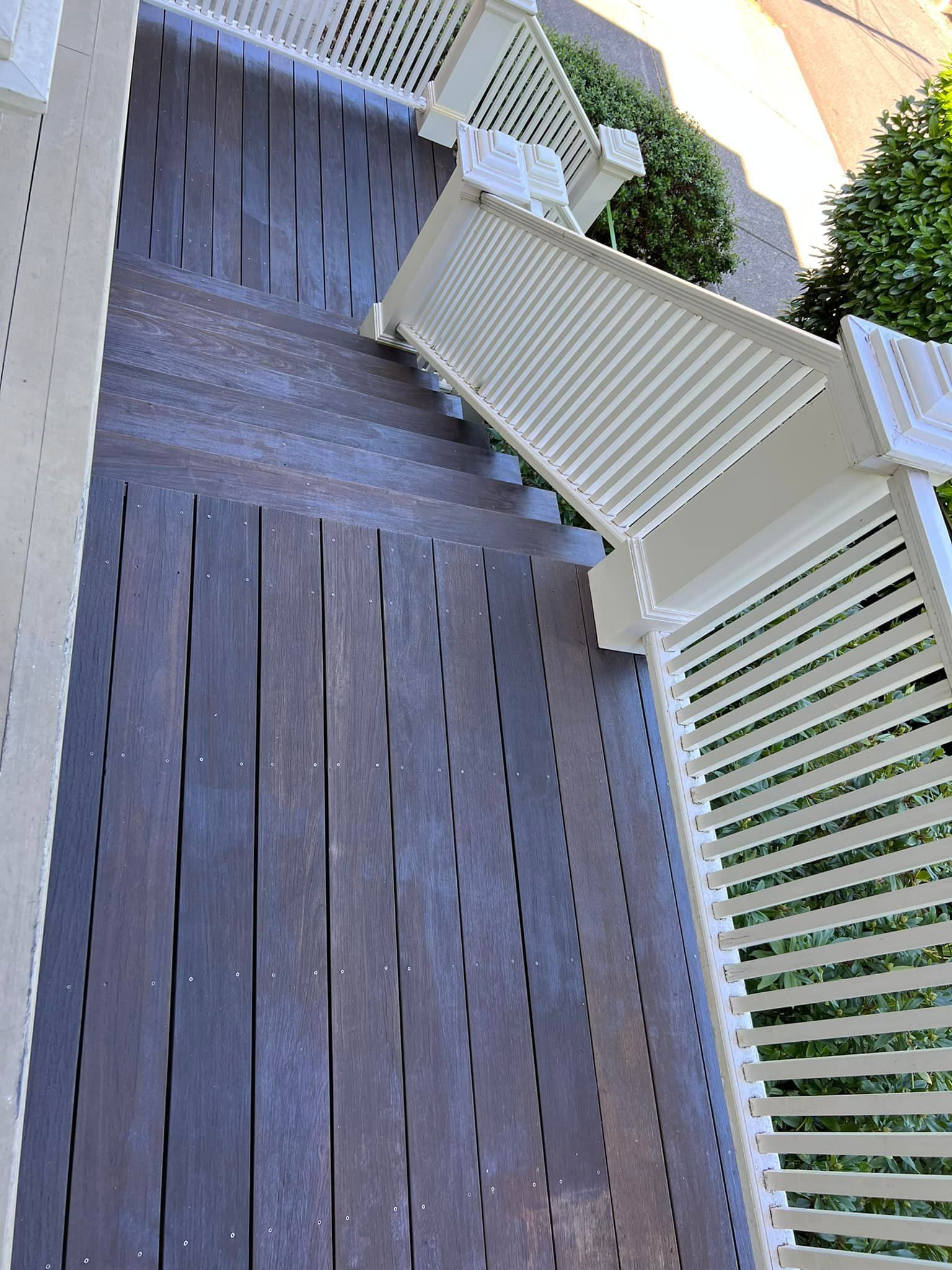 A high-angle view looking down a set of dark brown wooden stairs with white railings, flanked by green bushes.