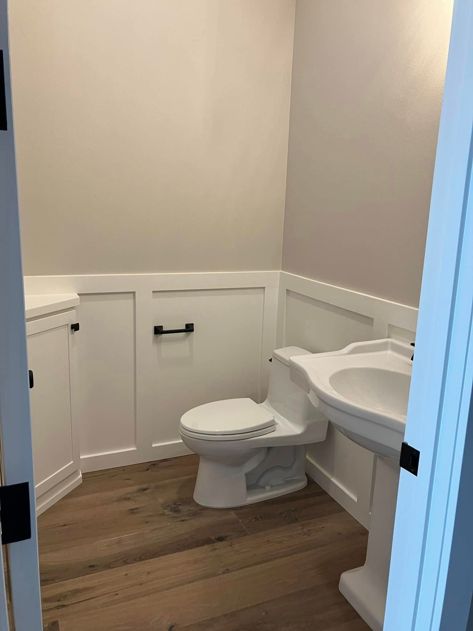 A small powder room with a pedestal sink, white toilet, light-colored walls, white wainscoting, and wood-tone flooring.