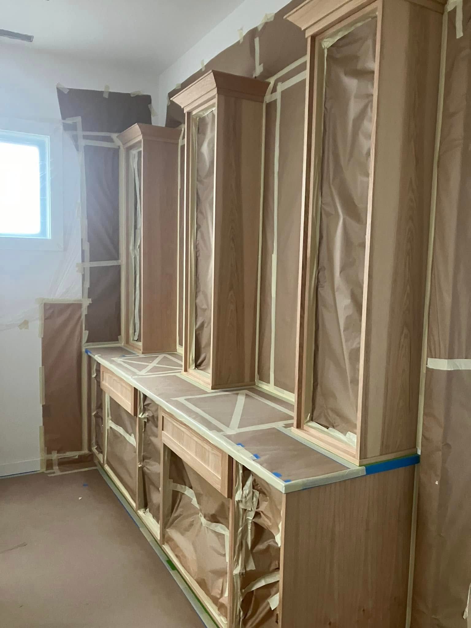 Kitchen cabinets being prepped for painting, covered in brown masking paper and painter's tape to protect surfaces.
