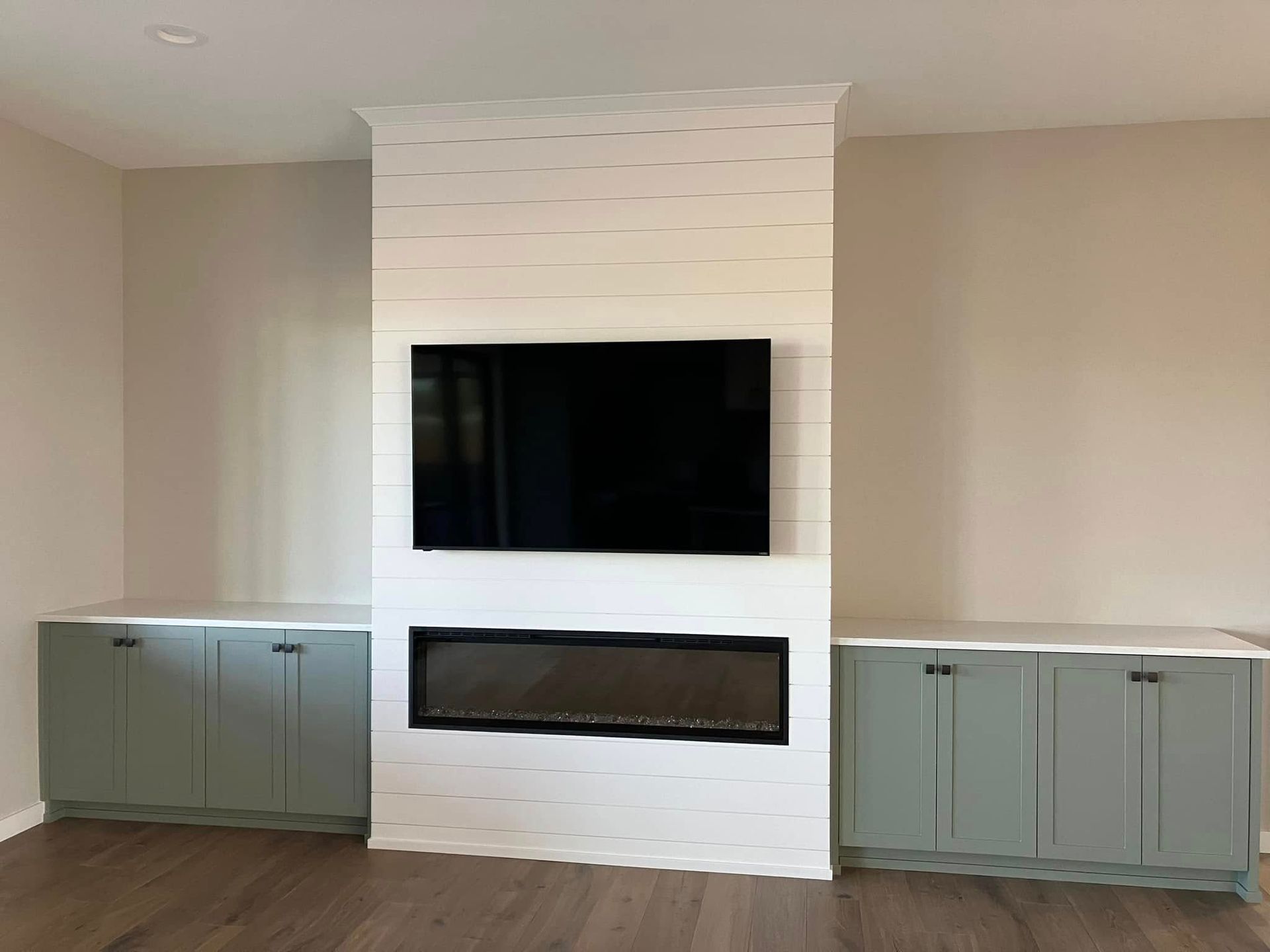A white shiplap fireplace wall with a built-in TV and fireplace, flanked by sage green cabinets on a wood floor.