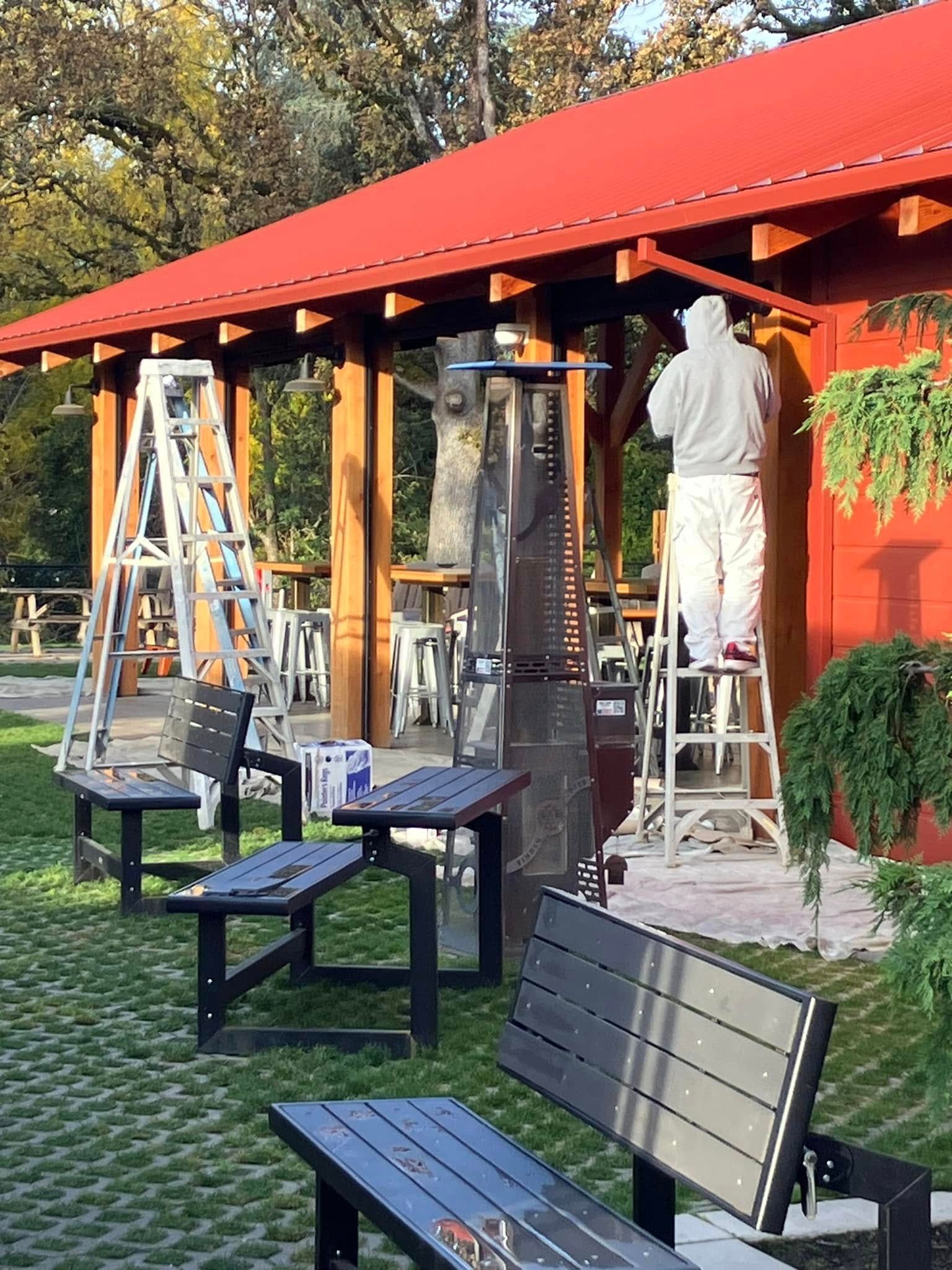 A person in protective coveralls works on the wall of a red-roofed building, near a ladder, outdoor benches, and a heater.