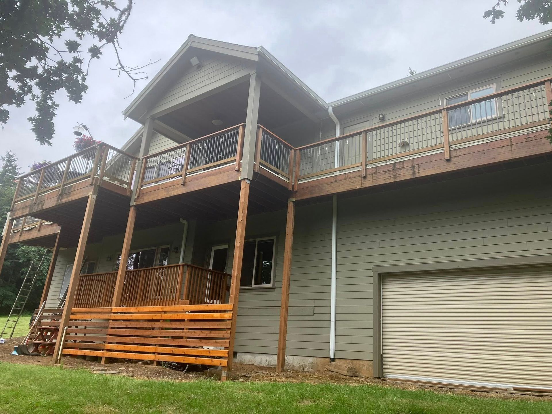 A two-story green house with a large wooden deck and balcony under a cloudy sky.
