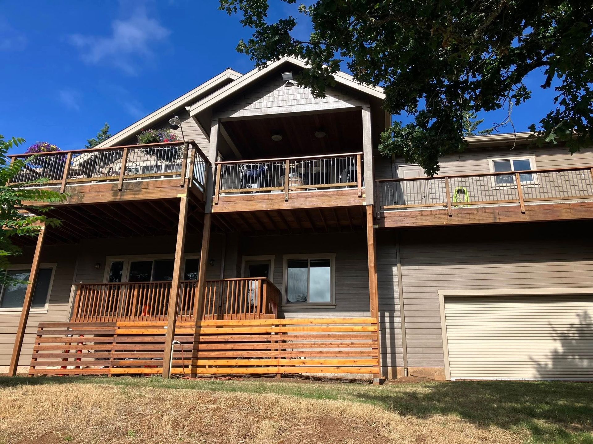 A two-story tan house with a large wooden deck, a covered balcony, and a garage under a clear blue sky.