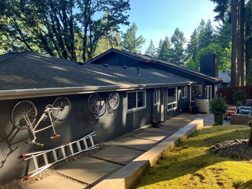 A dark grey house exterior with two mounted bicycles and a ladder on the wall, set along a paved path in a wooded area.