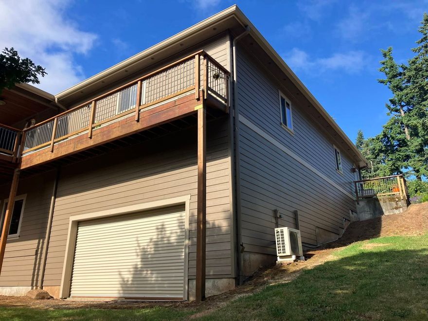 A two-story house with dark grey siding, a wooden deck, a roll-up garage door, and an external AC unit on a grassy slope.