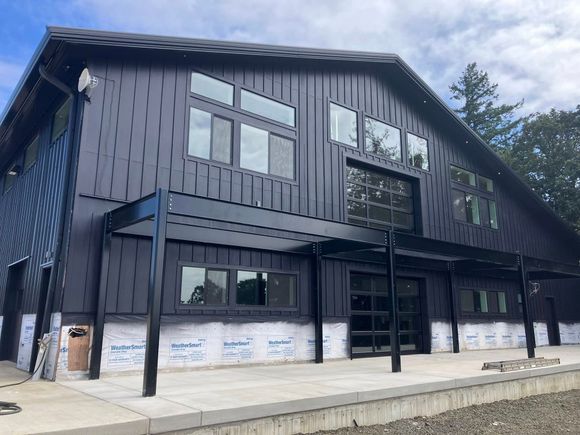 A modern, dark-sided building under construction with exposed metal beams, large windows, and a concrete patio.