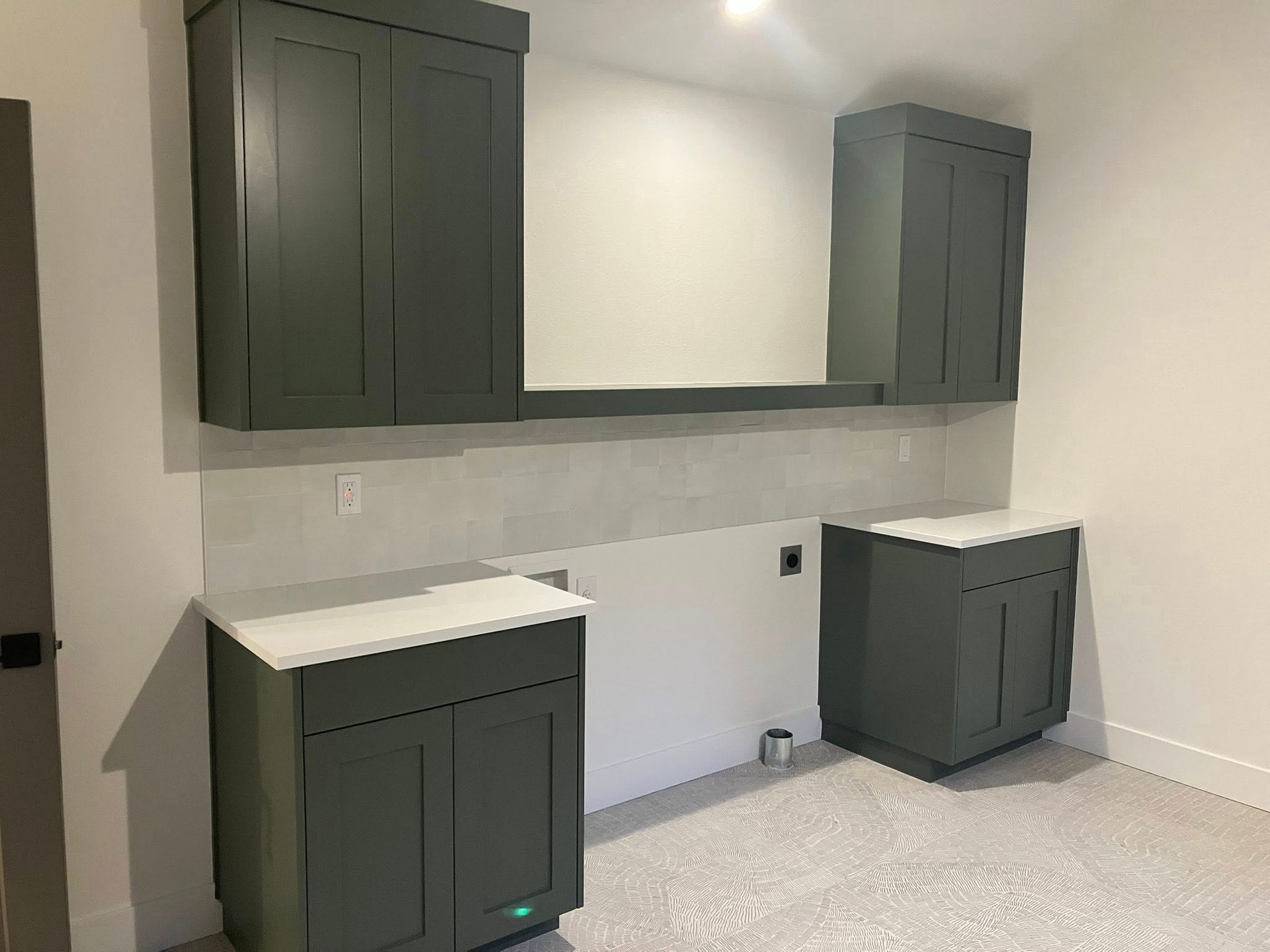 Dark green cabinets and white countertops in a laundry room with a light gray tiled backsplash and patterned tile floor.