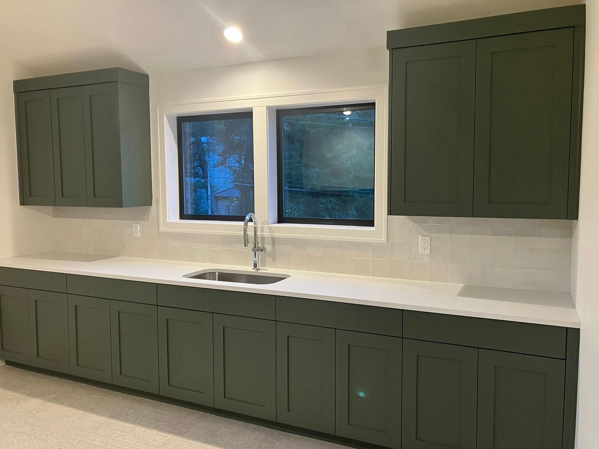 A kitchen with olive green cabinets, white countertops, and a double-basin sink positioned beneath two windows.