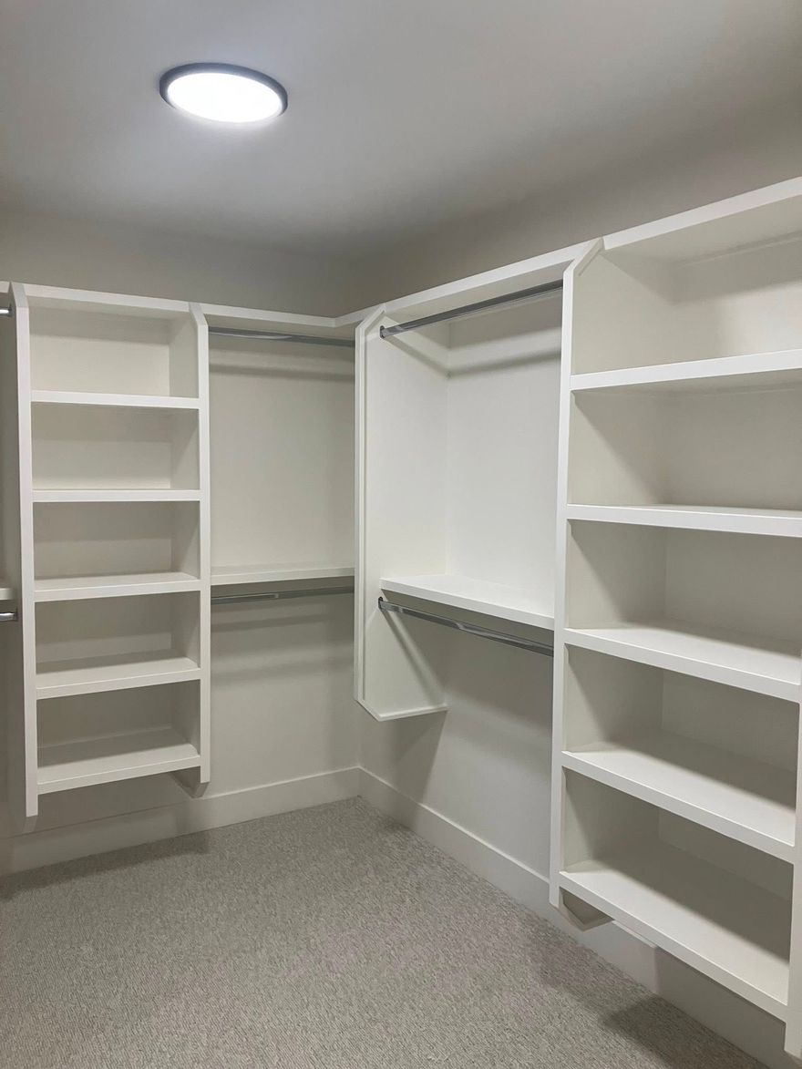 An empty walk-in closet with white shelving units, hanging rods, and gray carpeted flooring.