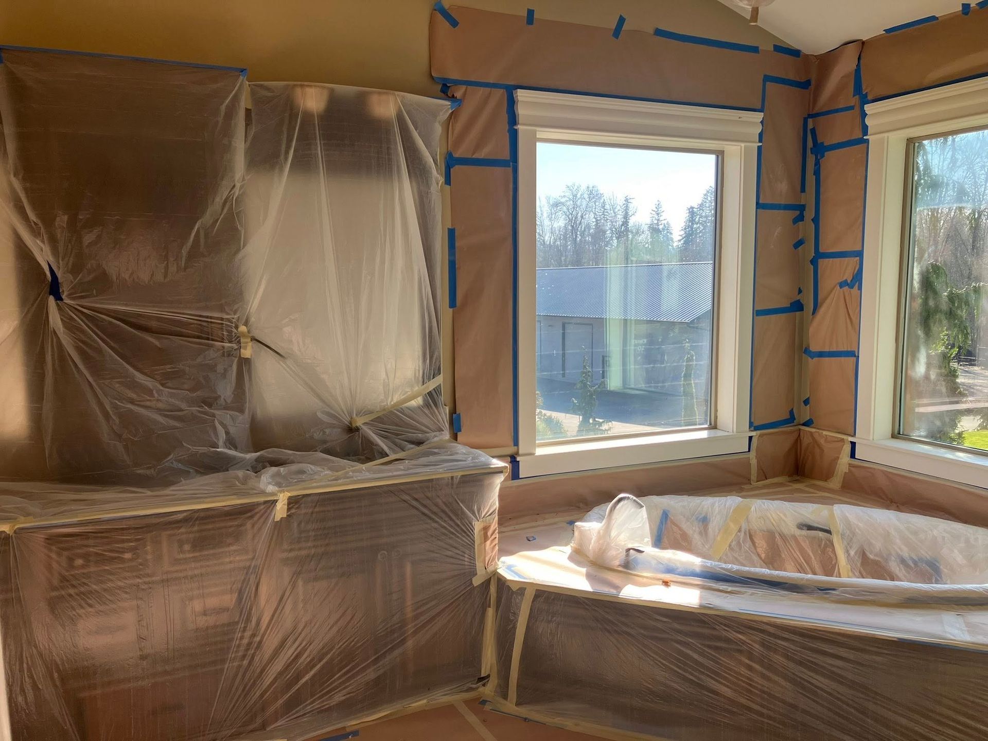 A bathroom renovation in progress with plastic sheeting and paper covering cabinetry, windows, and a bathtub.