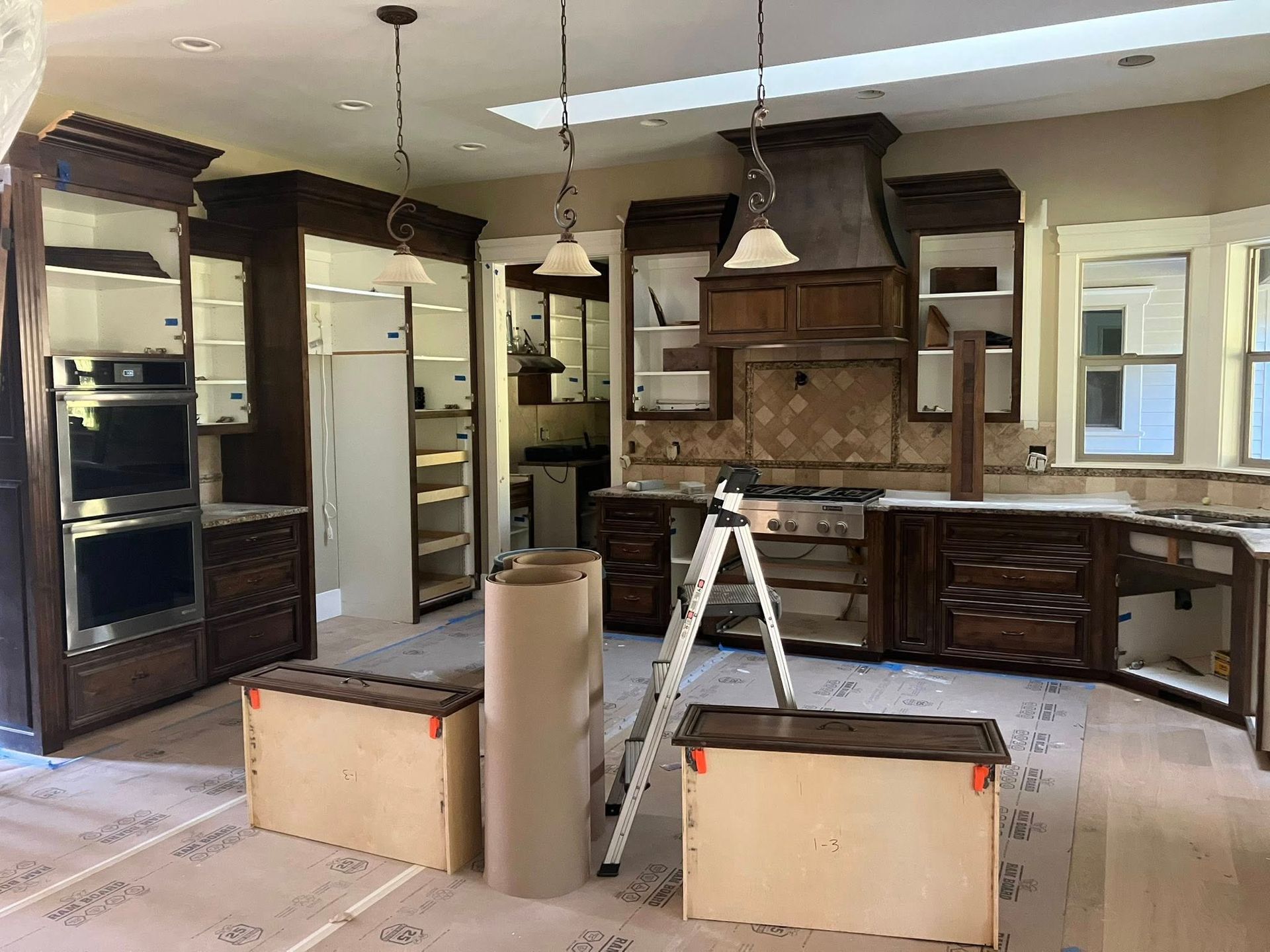 A kitchen undergoing renovation with dark wood cabinets, a ladder, and drawer fronts placed on a paper-covered floor.