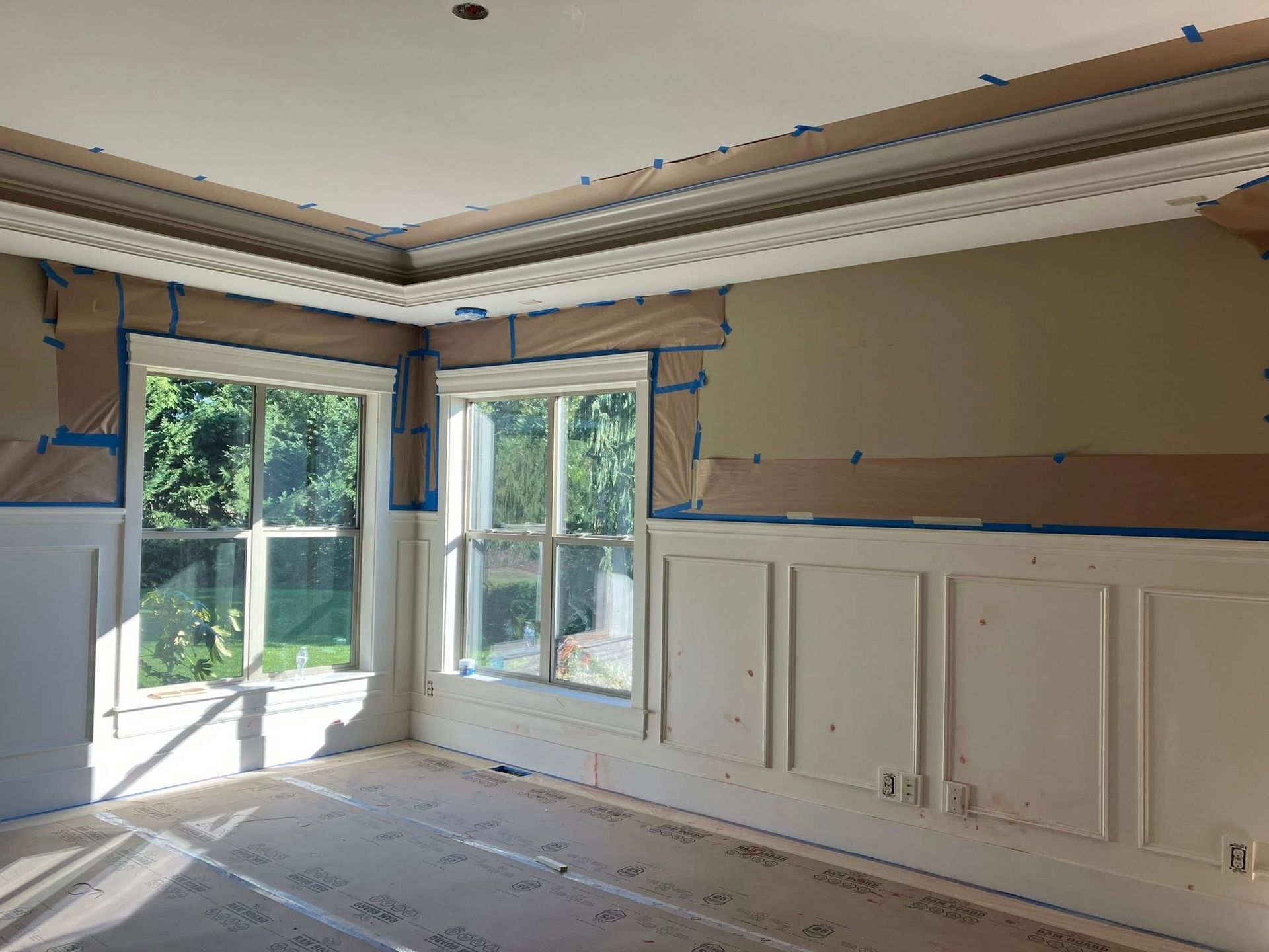 A room under renovation with white wainscoting, walls masked with brown paper, and ornate crown molding against a ceiling.