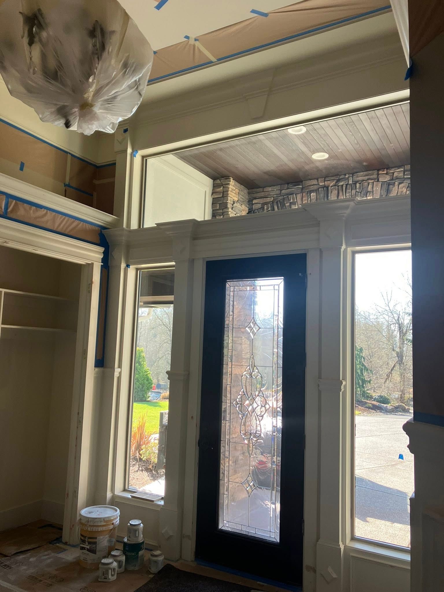 An interior view of an entryway with a dark, glass-paneled front door flanked by large windows, under renovation.