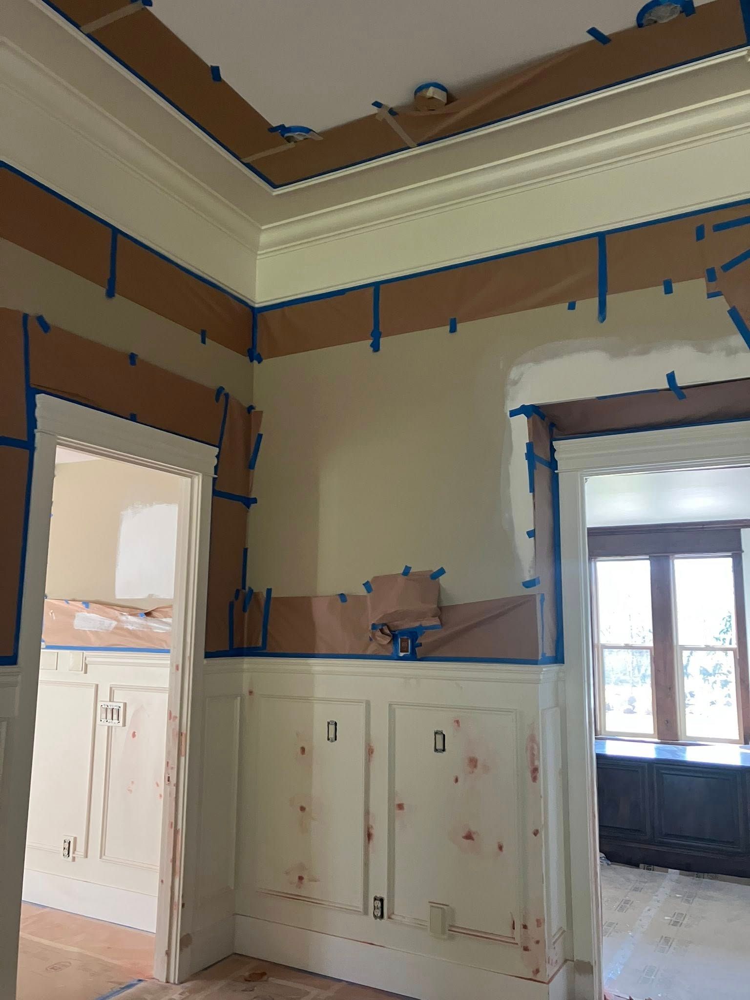 A room under renovation with beige walls, crown molding, and wainscoting, featuring blue painter's tape on all edges.