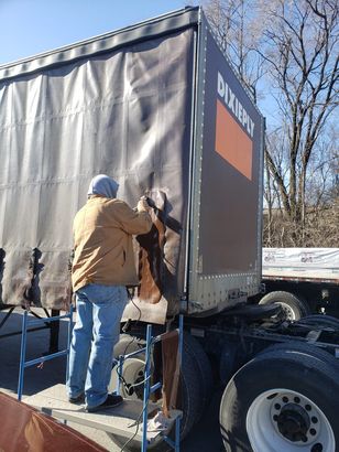 Man repairing a semi-trailer's side. He is wearing a brown jacket and blue jeans.