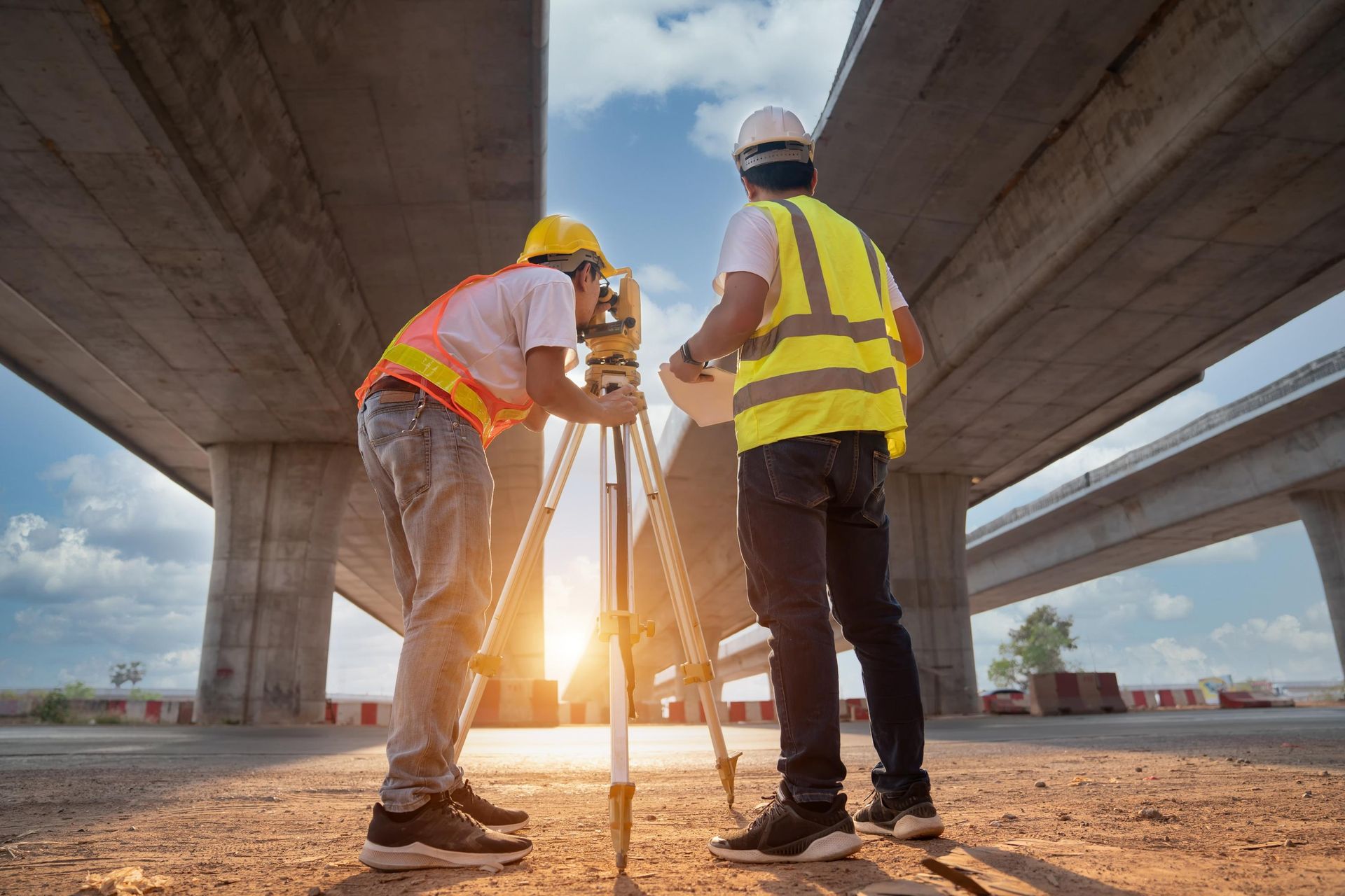 Two construction workers using surveying equipment under a highway overpass.