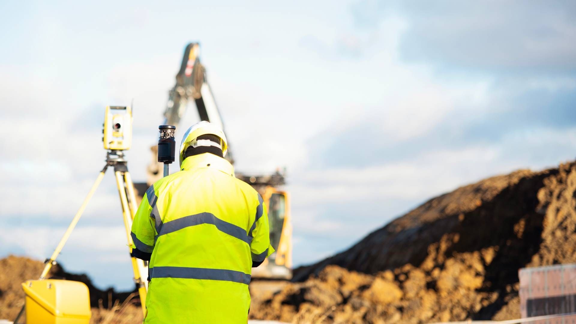 A surveyor in a high-visibility jacket uses a total station on a tripod at a construction site with heavy machinery.