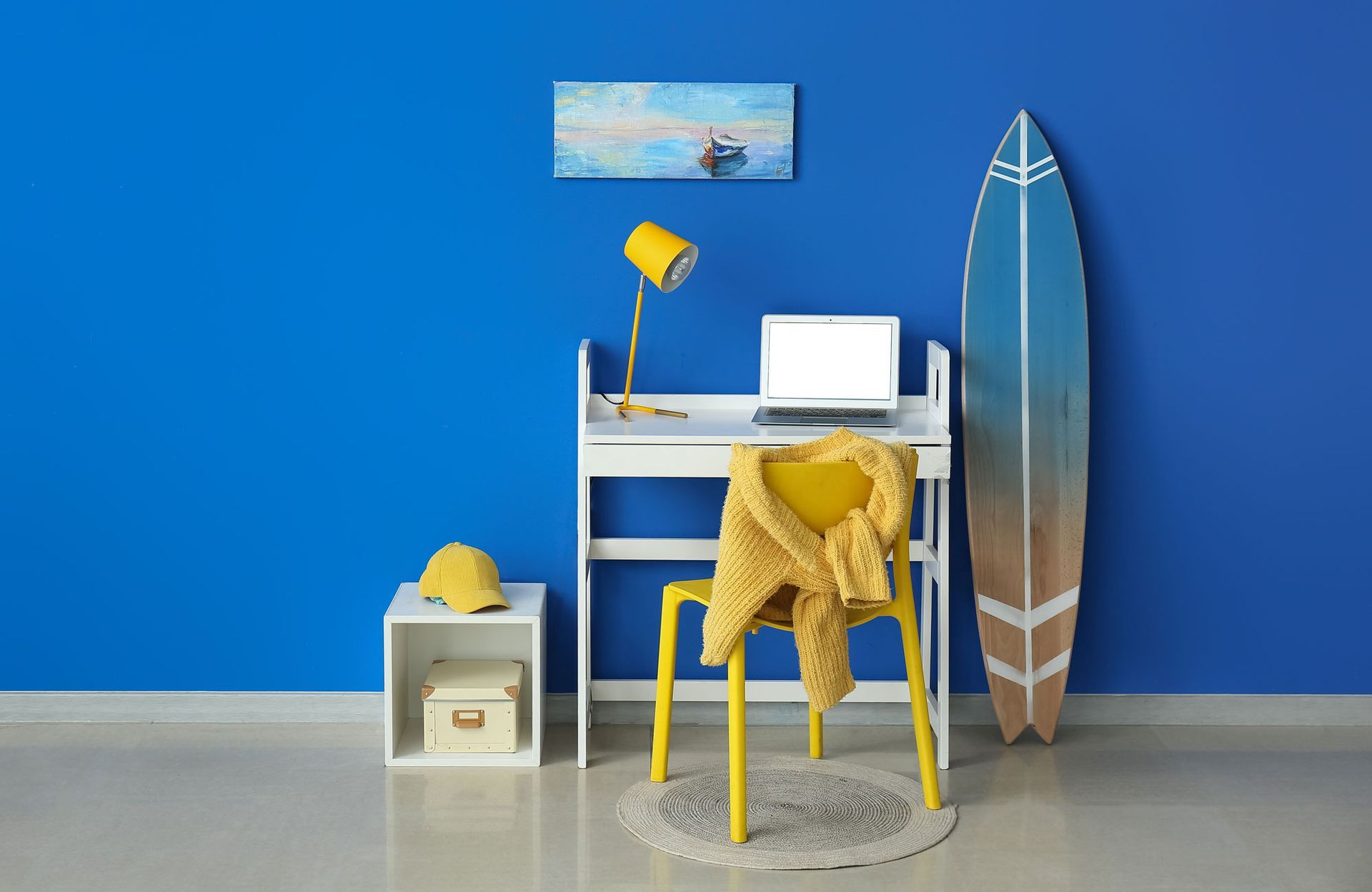 A blue room with a desk , chair , surfboard and laptop.