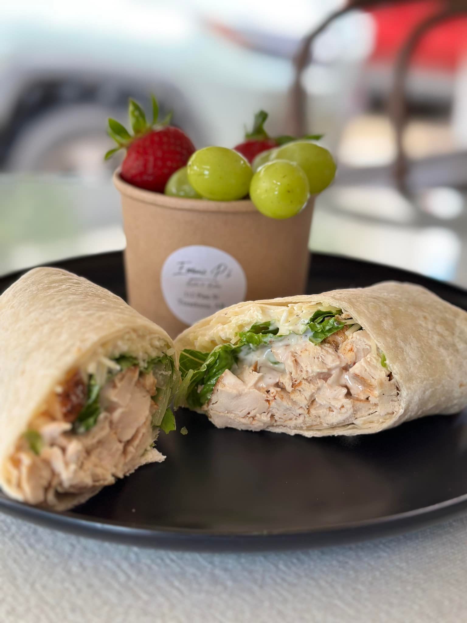 A chicken wrap on a black plate next to a cup of grapes and strawberries.