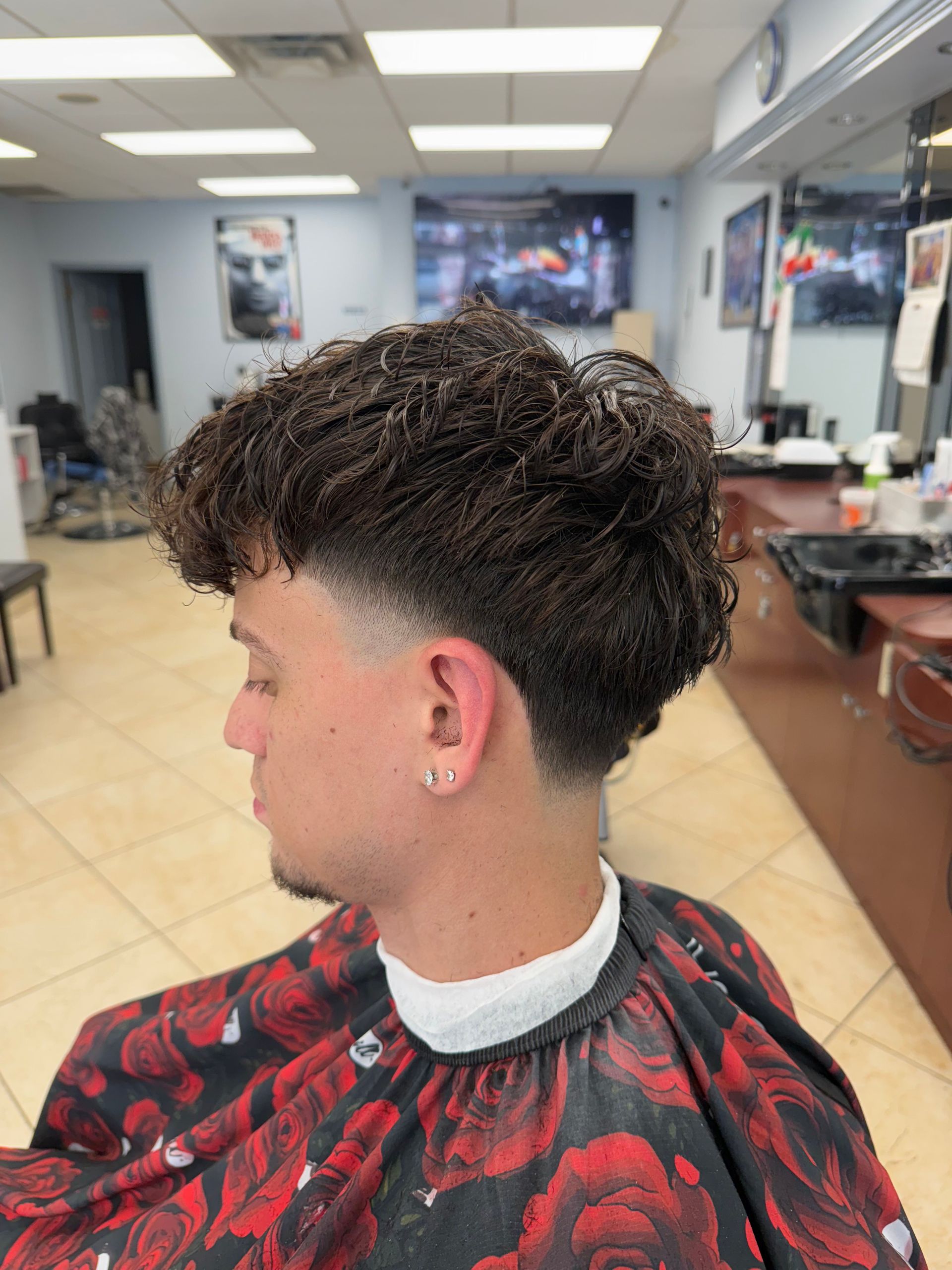 Man with wavy hair and a faded haircut in a barber shop; wearing a cape.