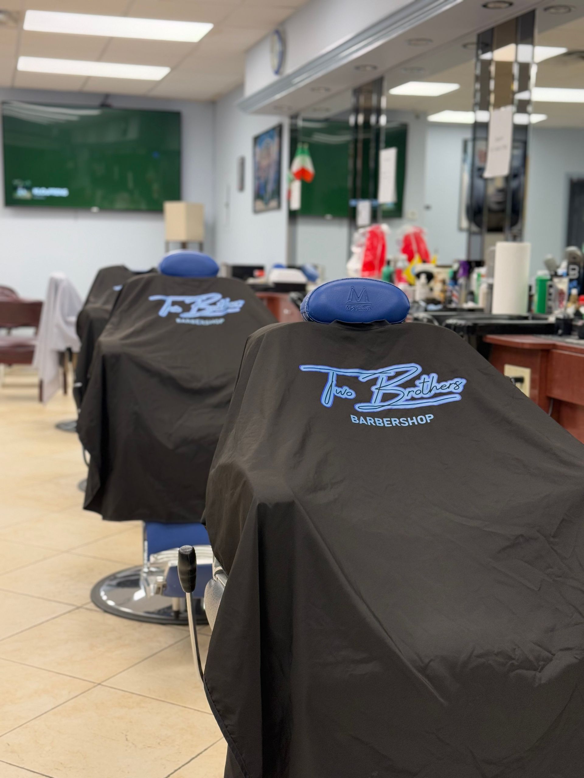 Two barber chairs covered in black capes in a barbershop. Blue headrests, logo on the capes.