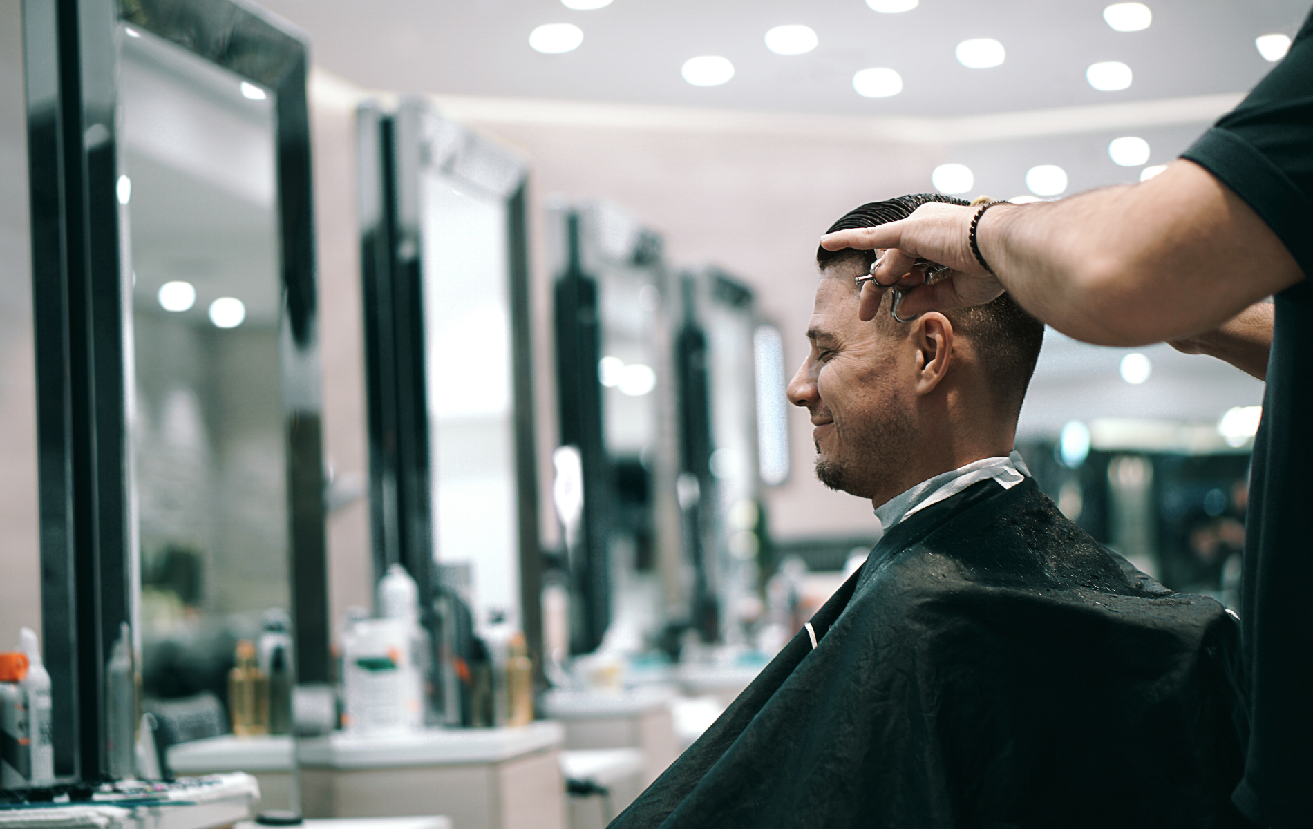 Person getting a haircut in a barbershop. Barber touching client's hair. Mirrors and shelves in the background.