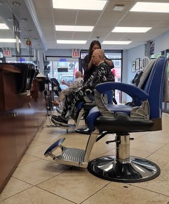 Barber shop scene: a person getting a haircut. Another person waits in a chair. Blue and black barber chairs, tiled floor.